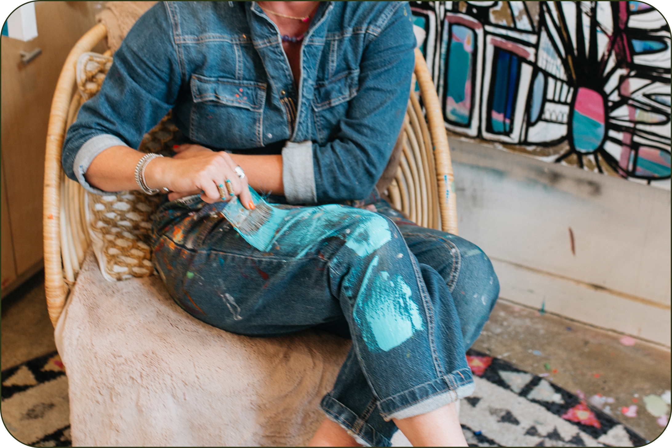A person sitting in a wicker chair, wearing a blue denim shirt and blue jeans with paint splatters, holding a paintbrush. A colorful abstract painting is visible in the background.
