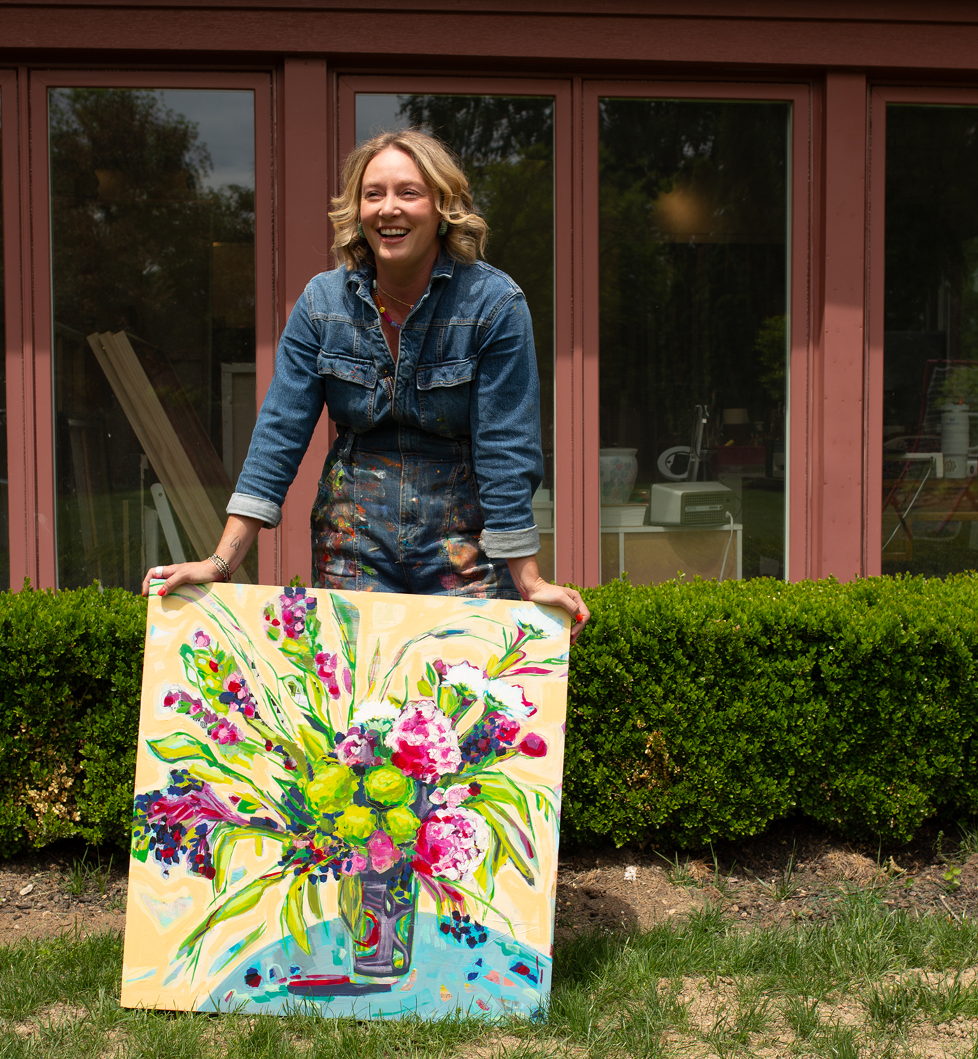 A smiling woman stands outdoors in front of a red-brown house and green hedges, holding a colorful painted canvas showing a bouquet of flowers in a vase.
