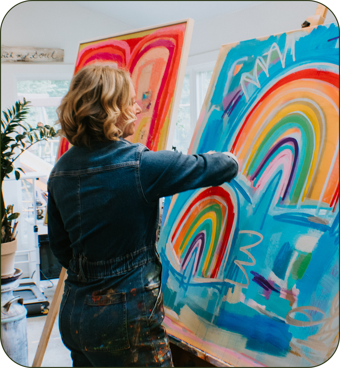 A woman in a blue denim jacket and paint-splattered overalls stands at an easel, painting a large colorful rainbow artwork on canvas in a bright studio.