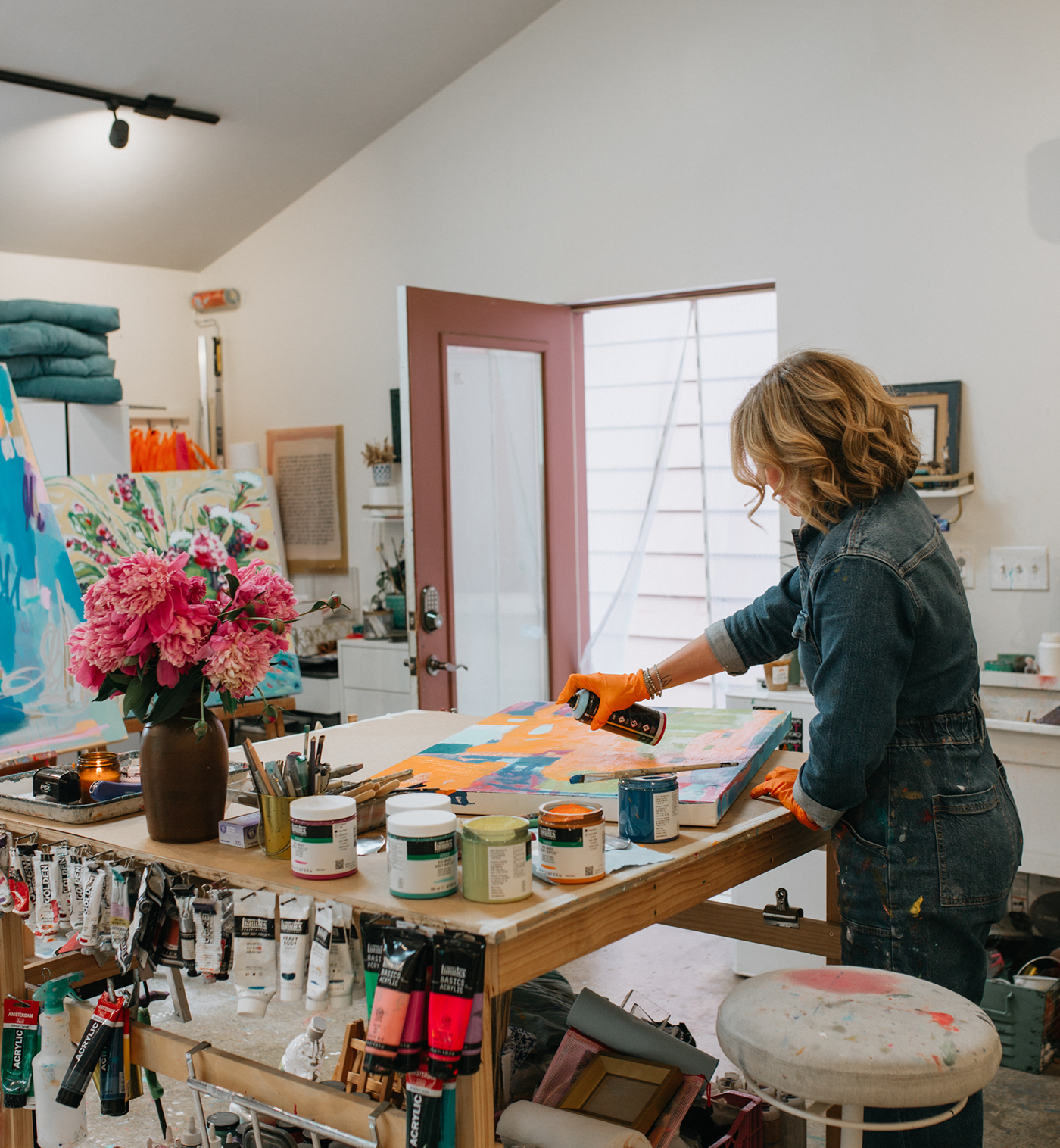 A woman in a denim jumpsuit and orange gloves stands at a wooden table in an artist's studio, working on a colorful painting. The table is covered with paint cans, brushes, and art supplies, with a vase of pink flowers in the foreground and a white wall in the background.