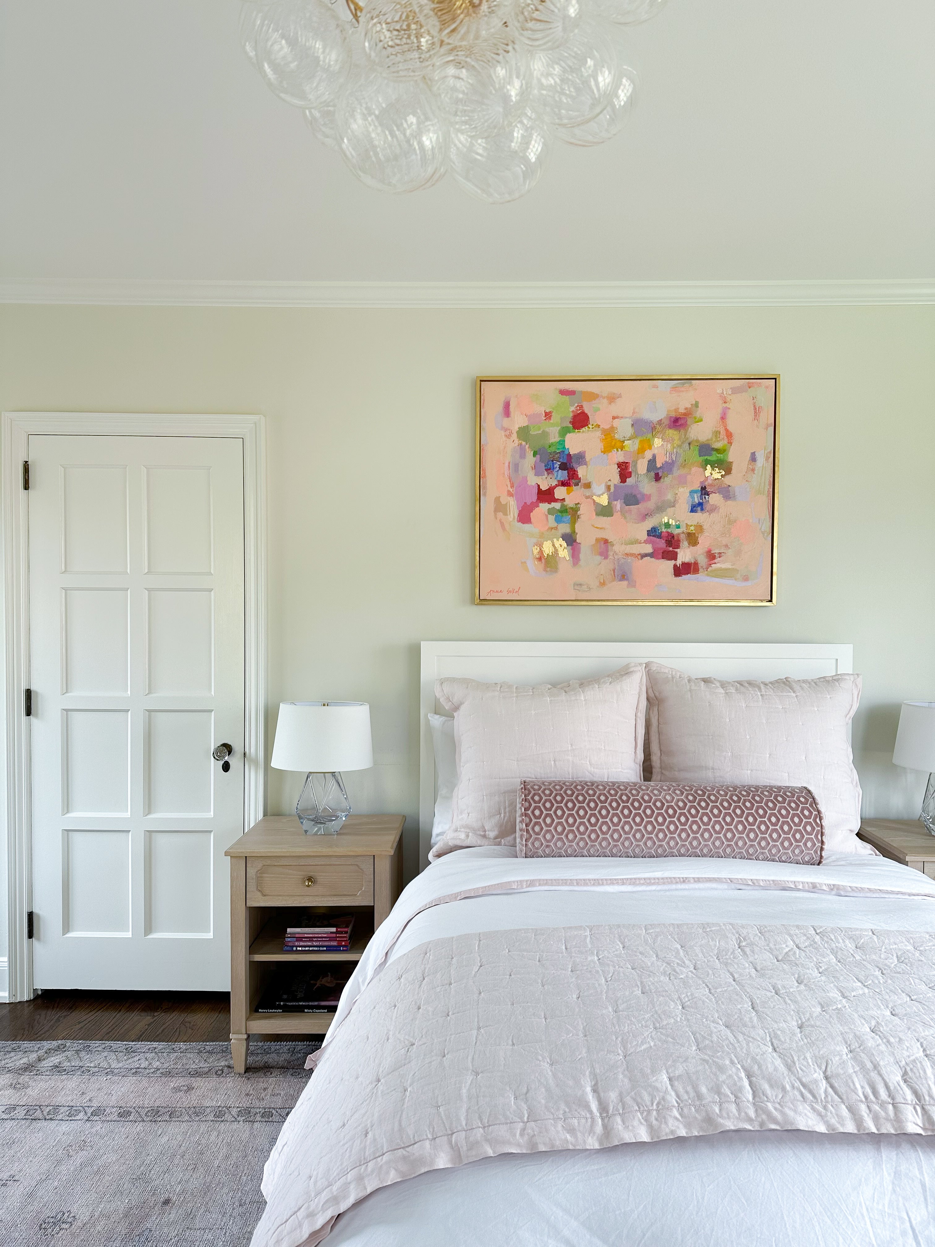 A styled bedroom interior featuring a bed dressed in white and blush bedding, a colorful abstract painting in a gold frame on the wall, a light wood nightstand with a lamp, and a white paneled door.