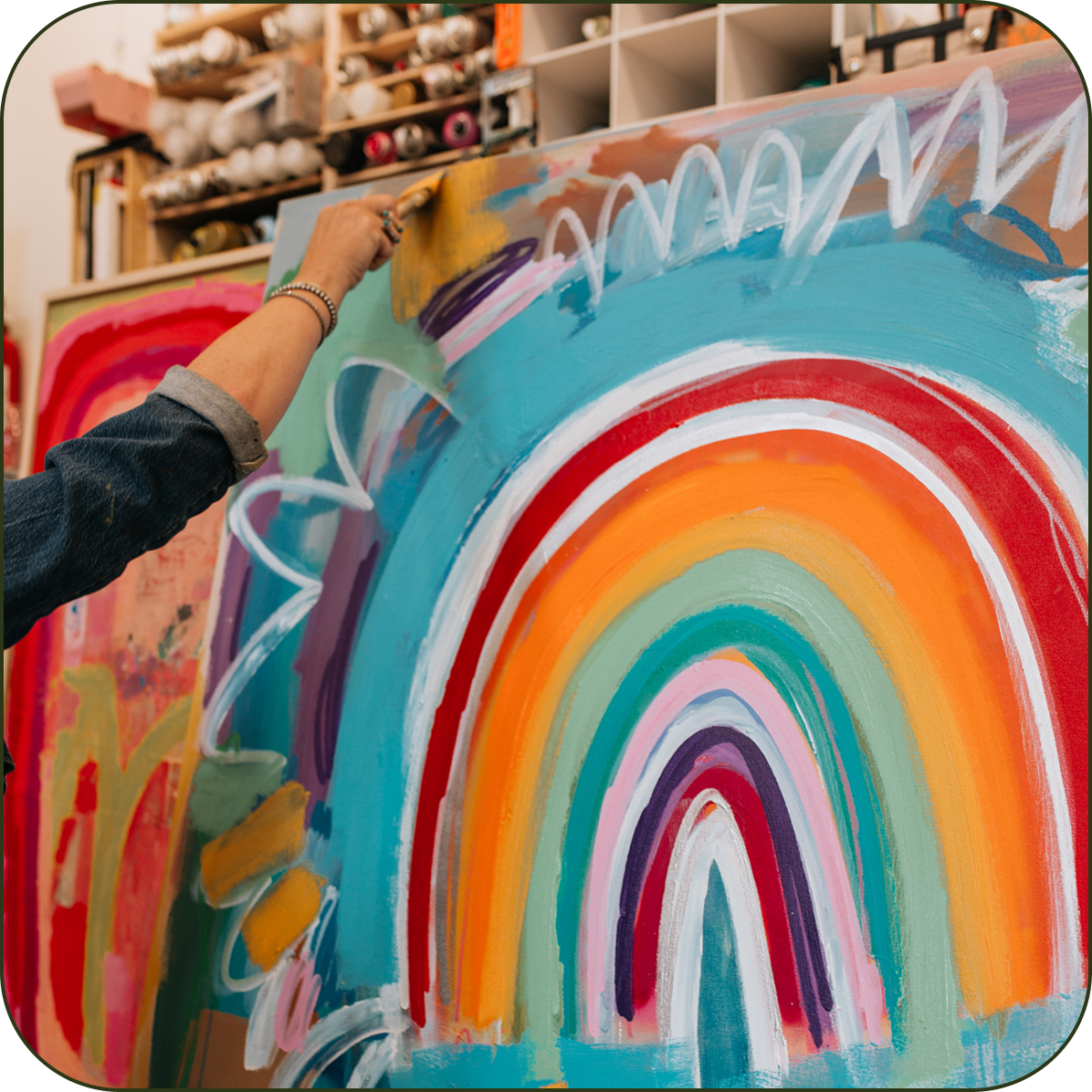 A person's arm and hand are shown painting a large canvas with a colorful abstract rainbow design in a studio setting.