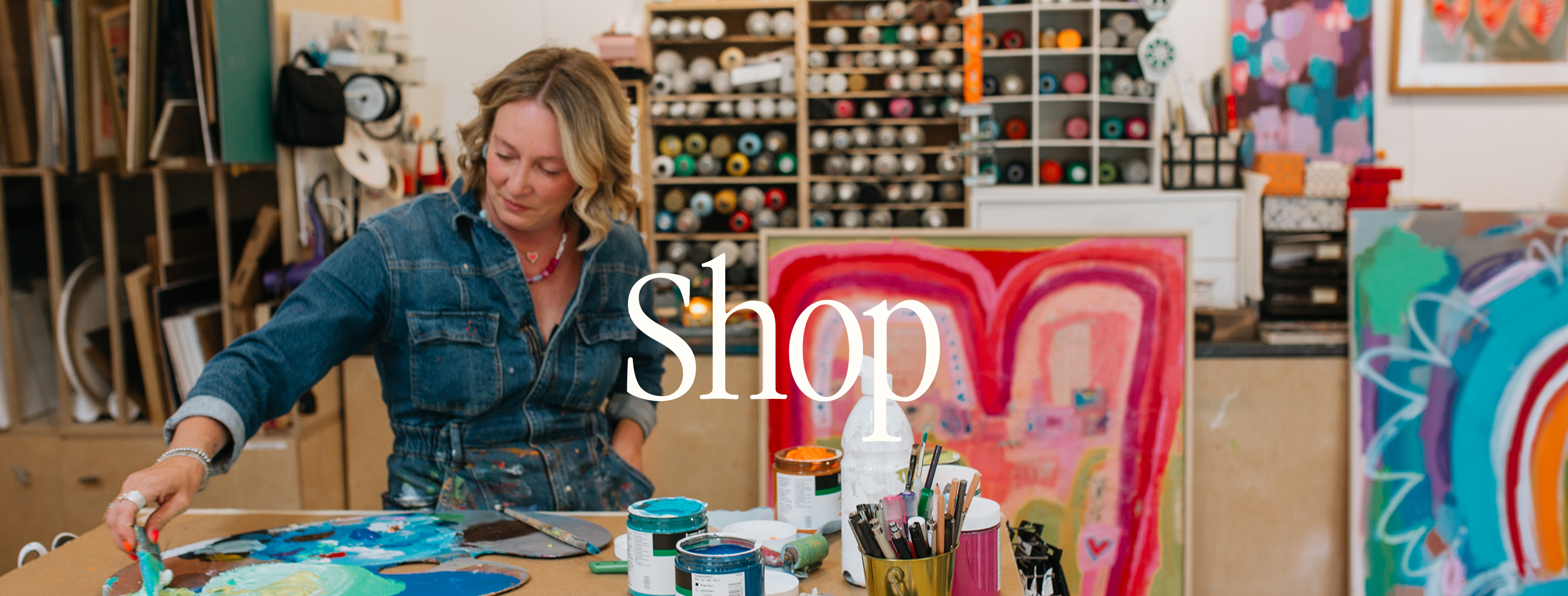 A woman in a denim shirt painting at a table in a colorful art studio, with a large white 'Shop' text overlay in the center.