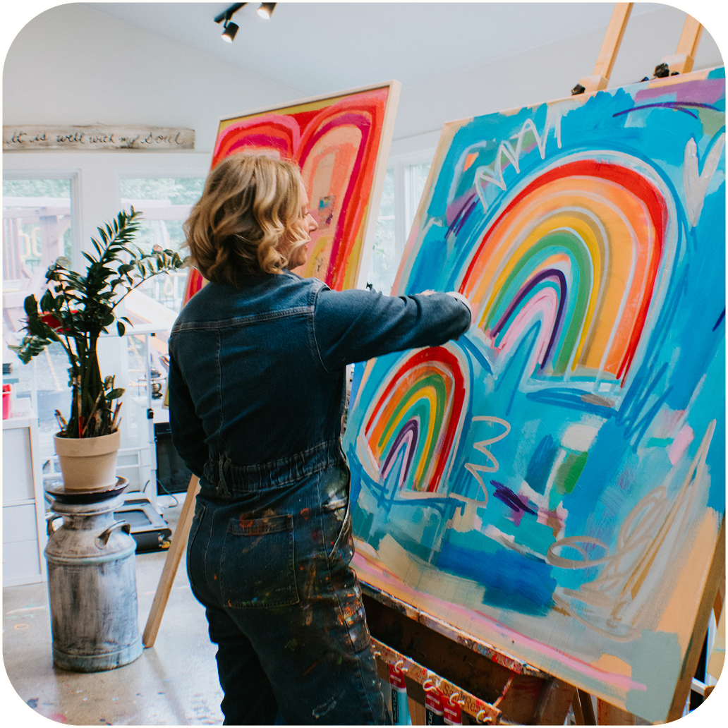 A woman in a blue denim jumpsuit stands at an easel painting a large colorful abstract rainbow artwork in a bright studio.