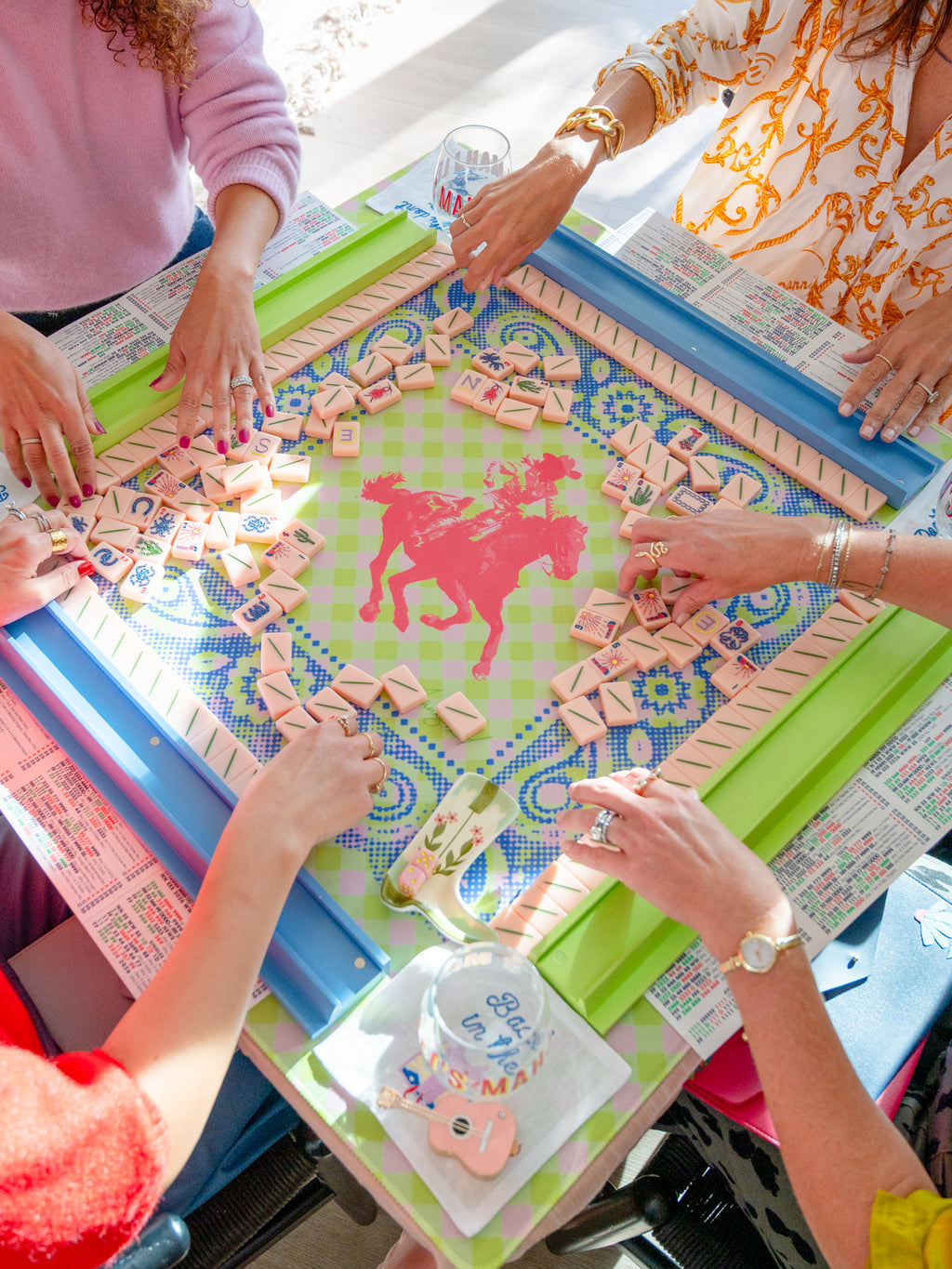 Several people are gathered around a colorful board game table, placing pink letter tiles onto a green and blue patterned game board with a red horse silhouette in the center.