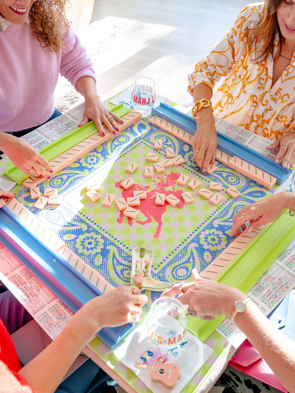 Several people are gathered around a colorful board game table, placing pink and beige game pieces on a patterned green and blue board. The scene is brightly lit with natural light, showing the hands of the players and parts of their clothing.
