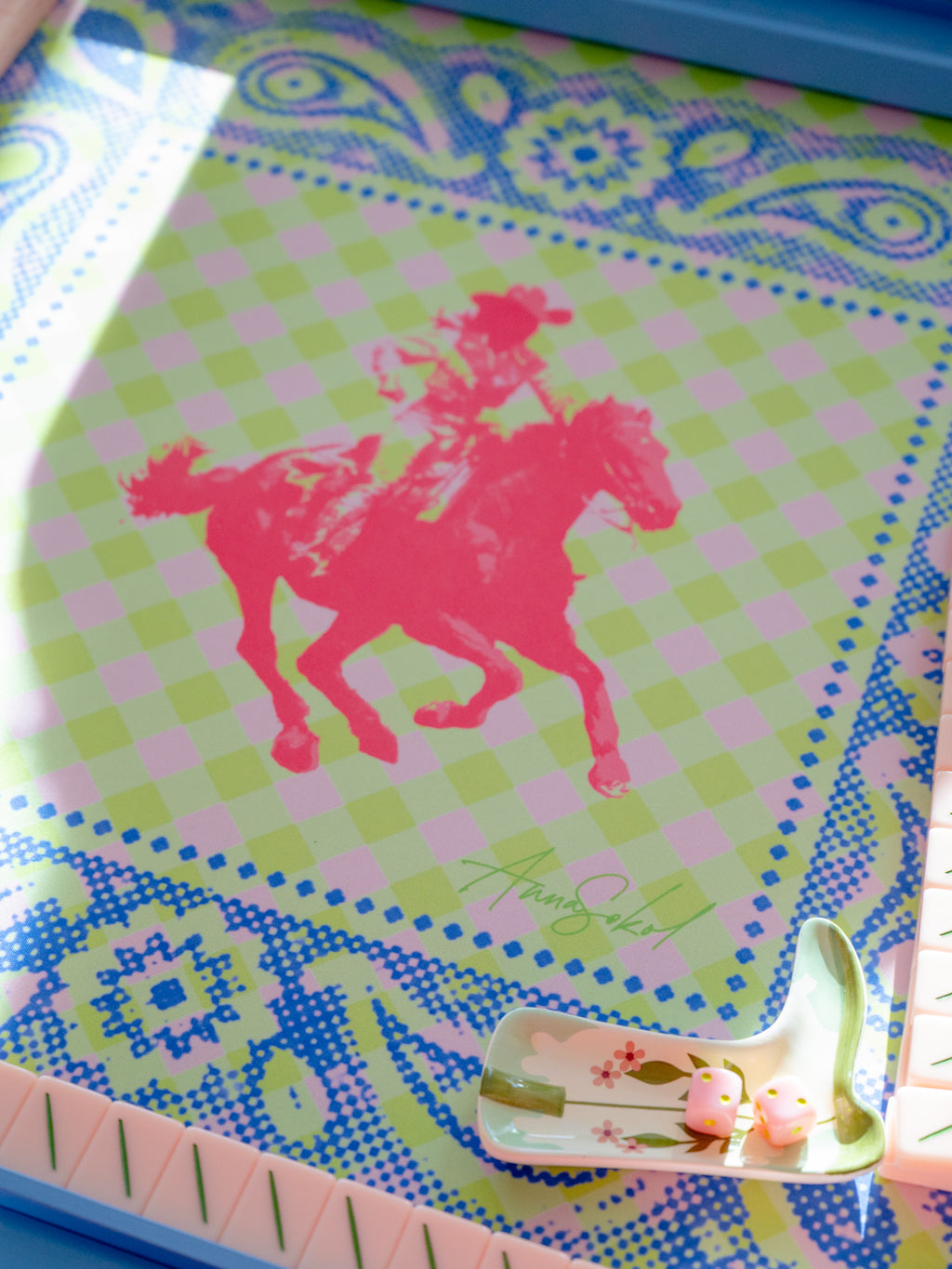 A close-up of a colorful printed surface featuring a pink silhouette of a rider on a horse over a pastel green and pink check pattern, surrounded by blue and green ornamental borders. A small decorative dish with pink duck figures sits in the lower right corner.