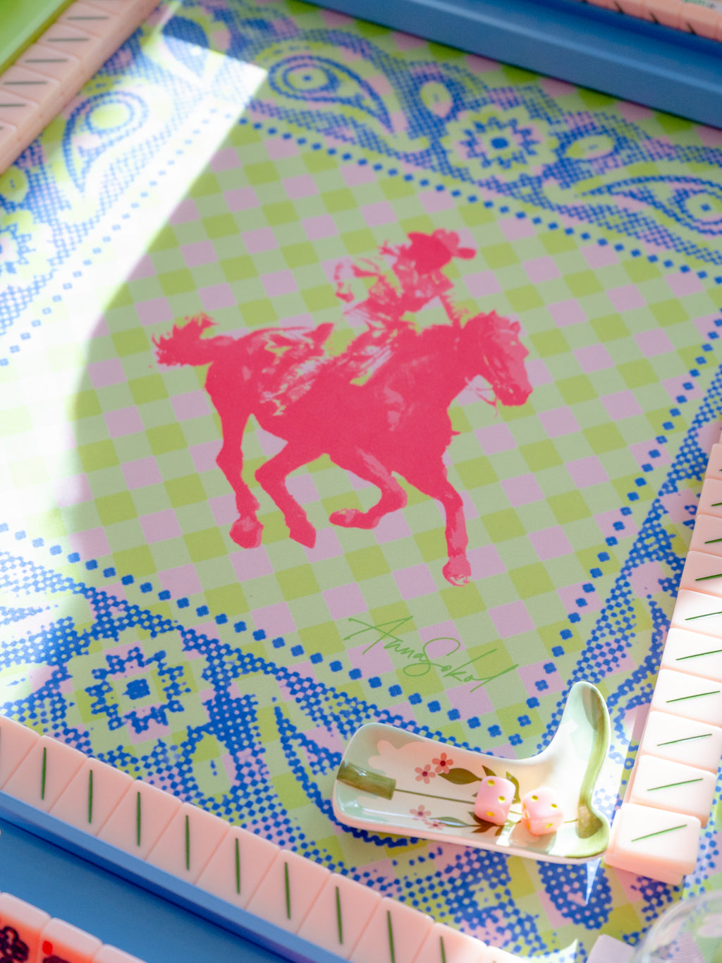 A close-up of a decorative tray or board featuring a pink silhouette of a cowboy riding a horse over a light green and pink checkered center, surrounded by a blue paisley border. A small white spoon with pink floral details rests on the lower right side.