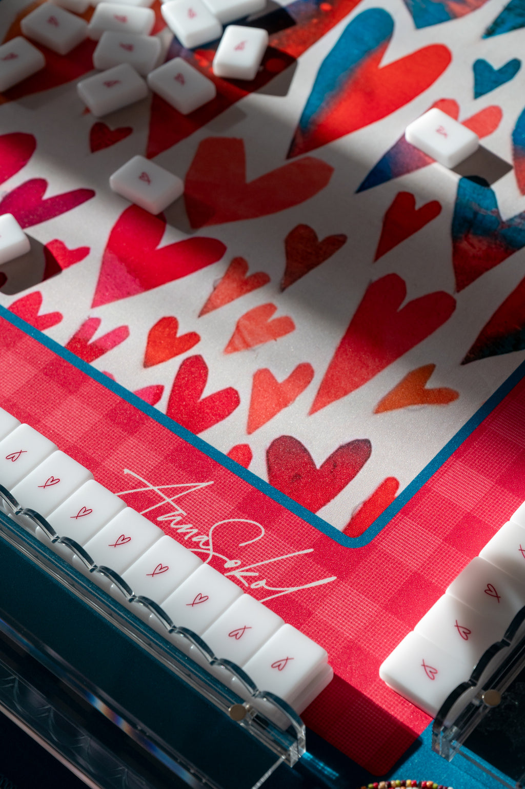 A close-up of a decorative game board featuring a red and white heart pattern, a red gingham border, and white square tiles with red hearts, photographed in bright sunlight with shadows.
