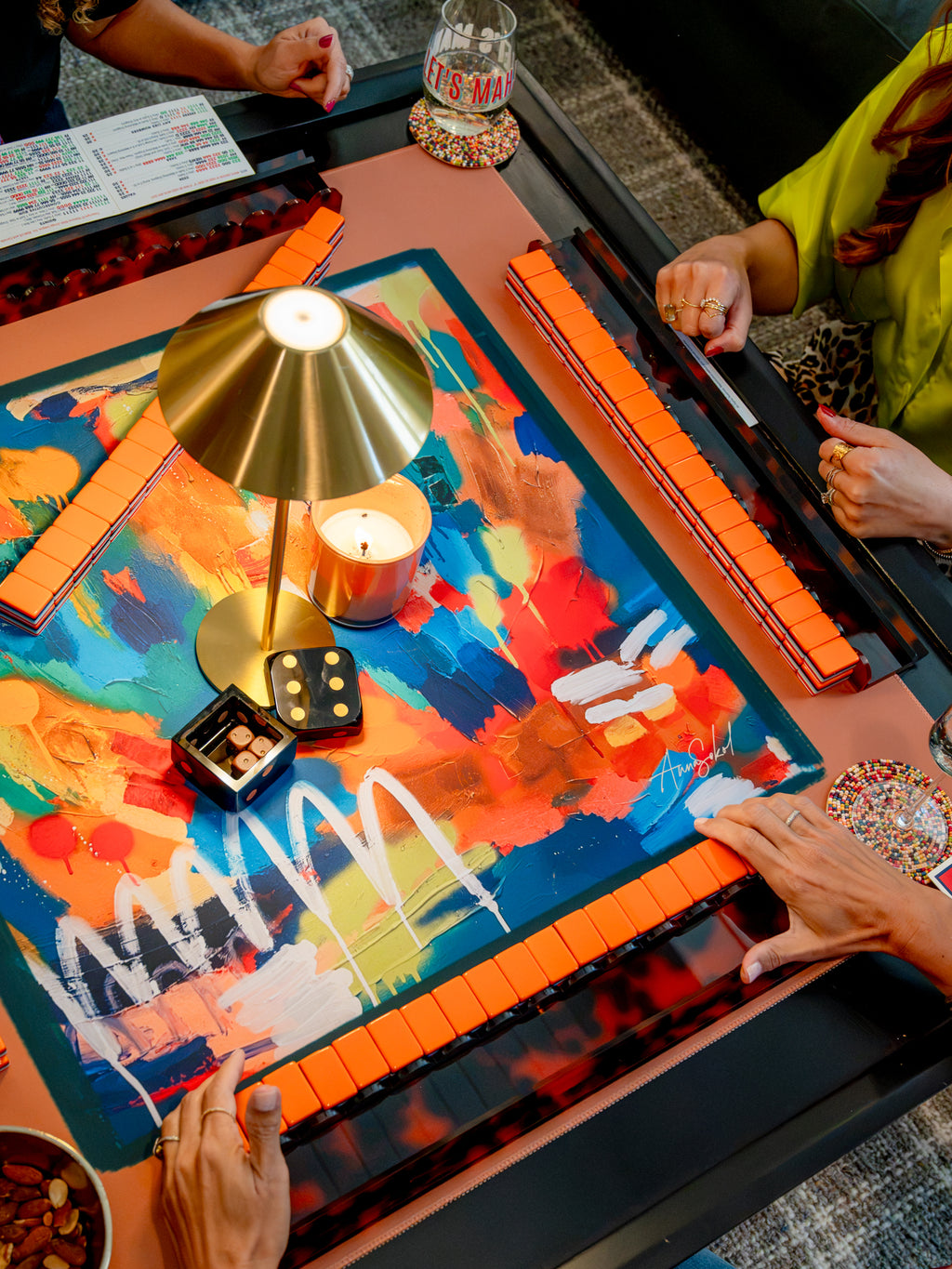 An overhead view of a social mahjong game in progress on a table, with multiple hands visible holding tiles. The table features a colorful abstract painted mat, a gold-colored dome lamp, a lit candle, dice, and a glass of water.