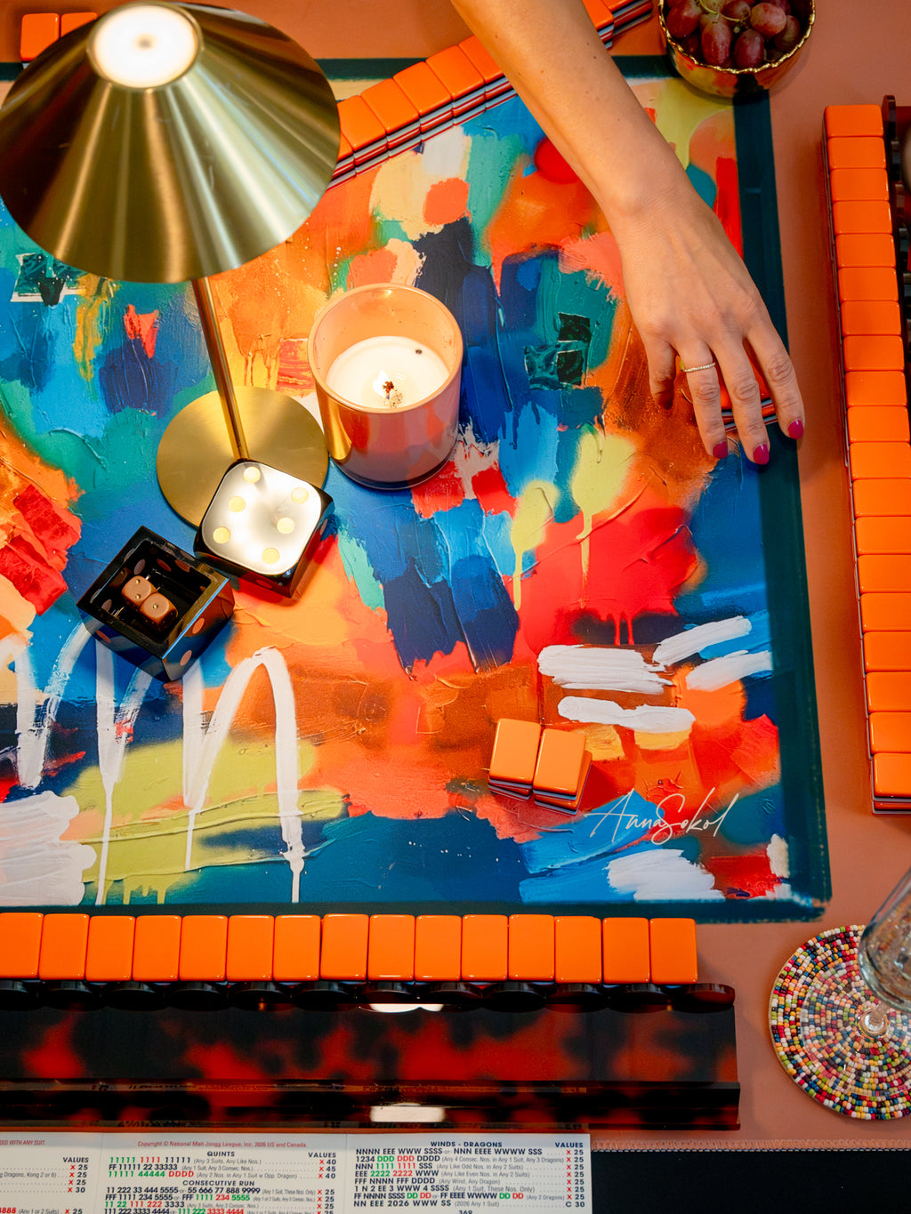 A top-down view of a colorful abstract art print or canvas on a table, surrounded by candles, a metallic lamp, and orange blocks. A person's hand with pink nail polish rests on the artwork. Anna Sokol signature at the bottom