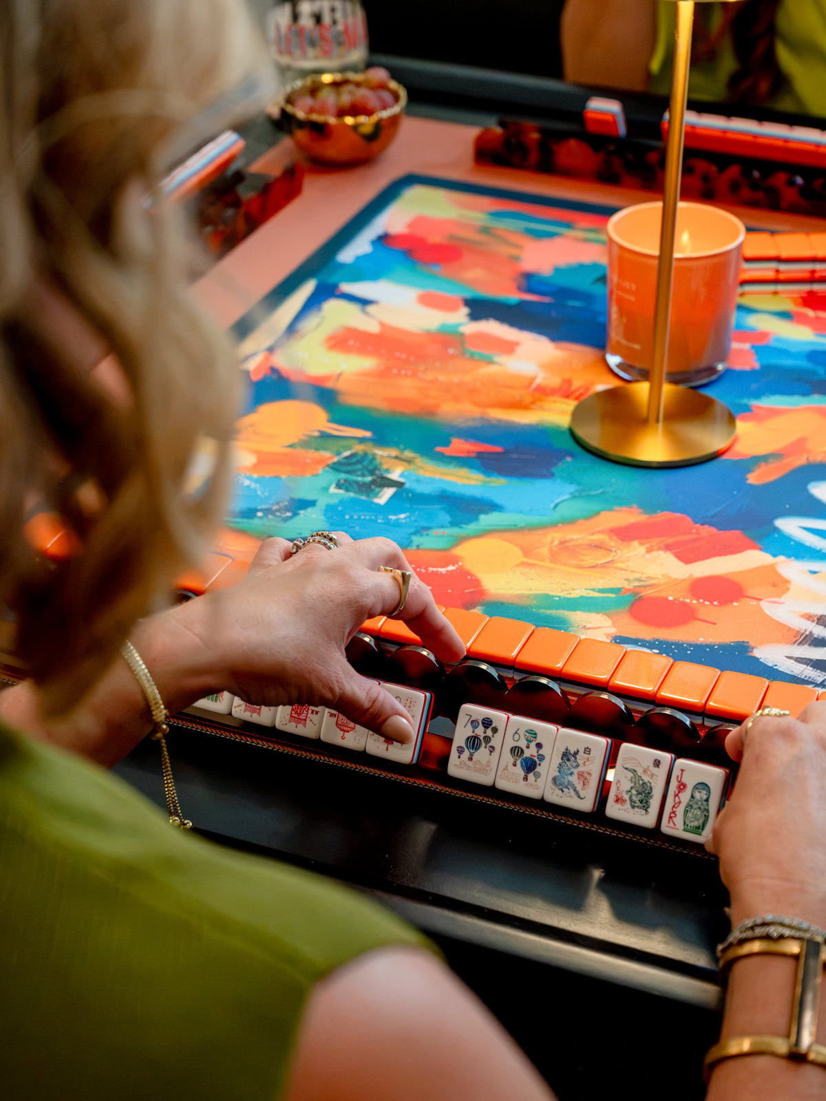 A close-up lifestyle shot of a person playing a colorful mahjong game, with their hands visible holding tiles over a vibrant, abstract-patterned board under warm ambient lighting. Anna Sokol signature at the bottom