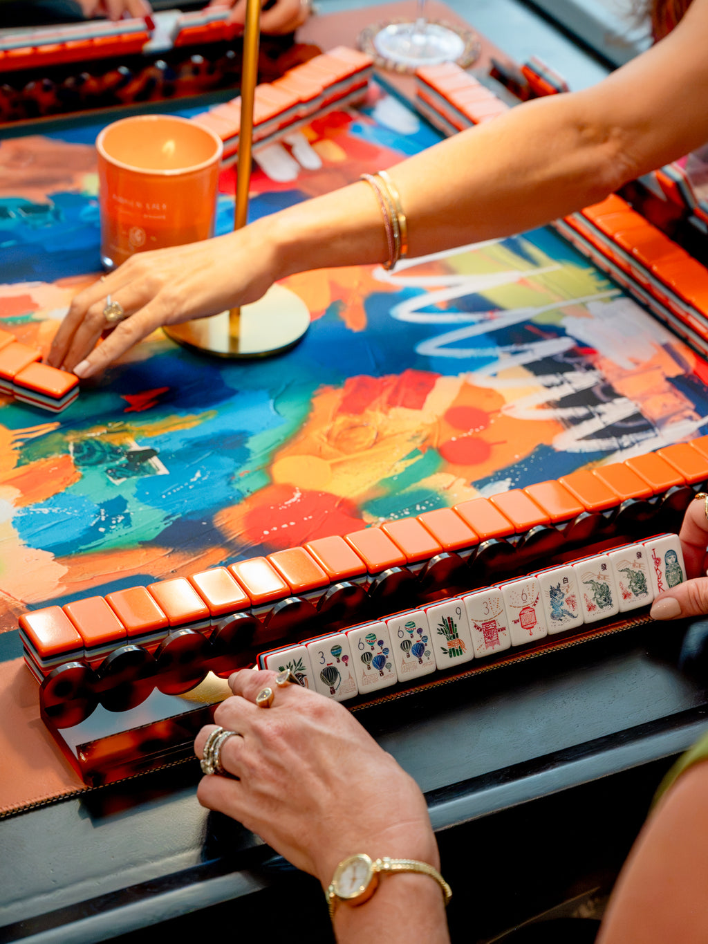 A close-up lifestyle shot of a colorful Mahjong game table in use, with a person's hands visible arranging tiles and interacting with the game. The table features a vibrant abstract multicolor design with orange, blue, yellow, and red tones, and the tiles have a glossy orange-red tortoiseshell finish. Anna Sokol signature at the bottom