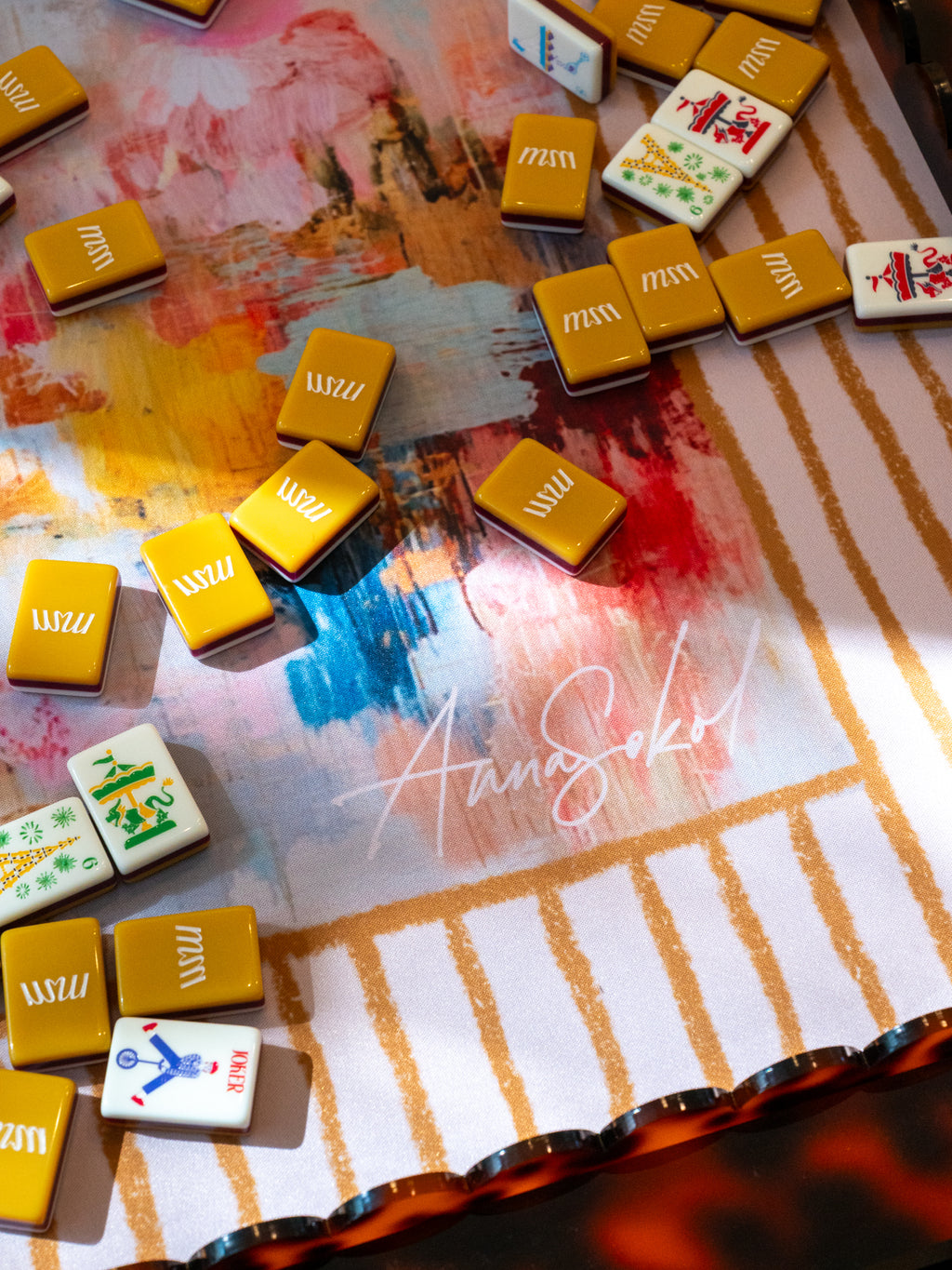 A close-up overhead view of mahjong tiles scattered on a decorative game mat with painted abstract brushstrokes and gold striped borders. Several tiles are visible, including yellow-backed tiles and white tiles with red, green, blue, and black markings. Anna Sokol signature
