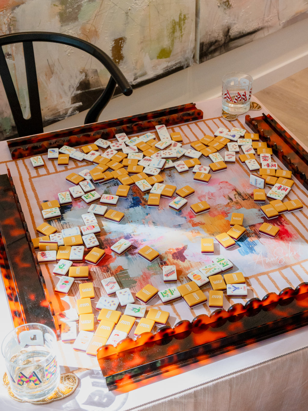 A decorative rectangular serving tray with a glossy tortoiseshell-style amber and black border, filled with many small white cards or tiles scattered across a painted abstract surface. The tray sits on a table in an indoor setting with a chair and wall art visible.