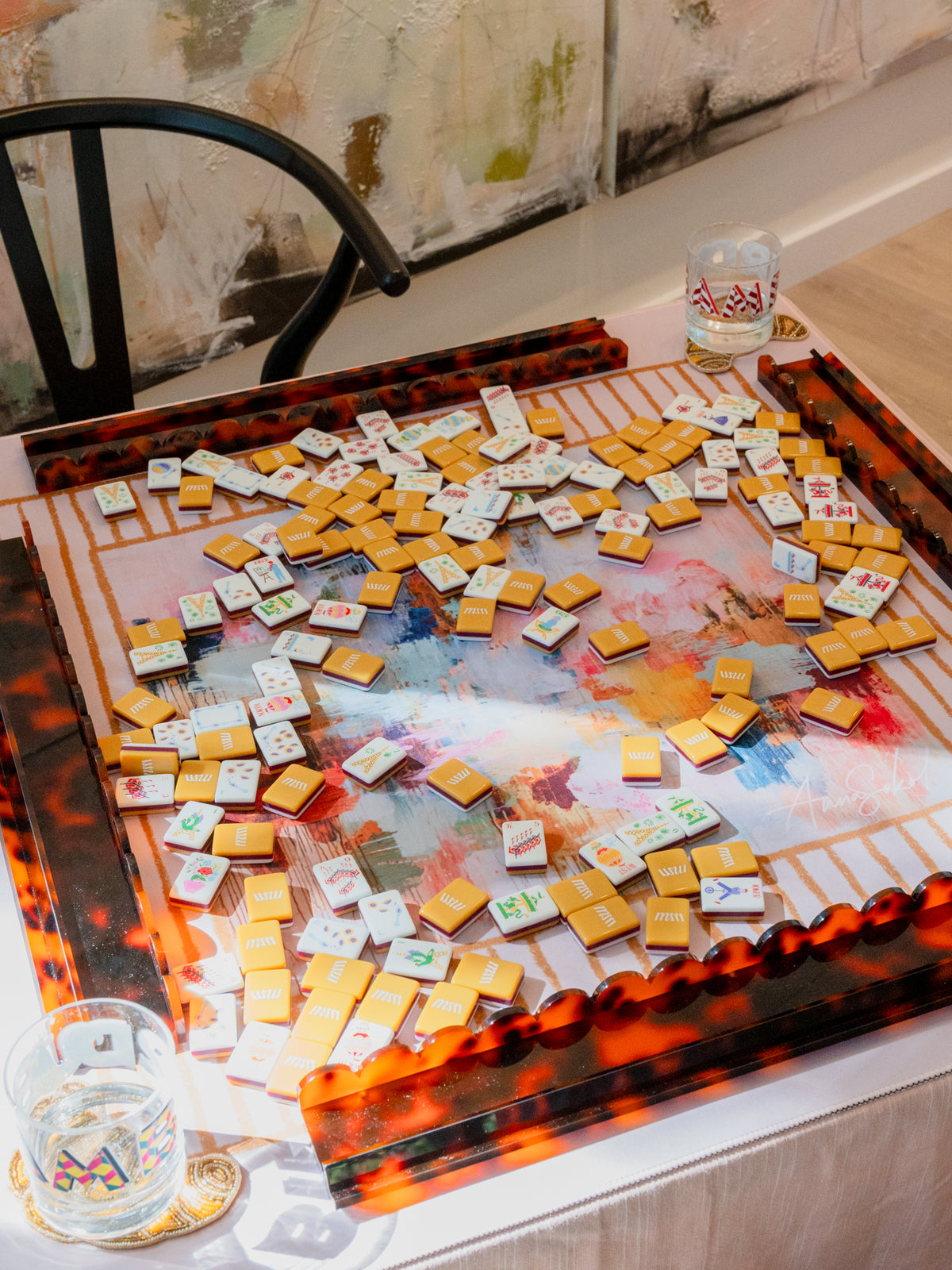 A decorative rectangular serving tray with a glossy tortoiseshell-style amber and black border, filled with many small white cards or tiles scattered across a painted abstract surface. The tray sits on a table in an indoor setting with a chair and wall art visible.