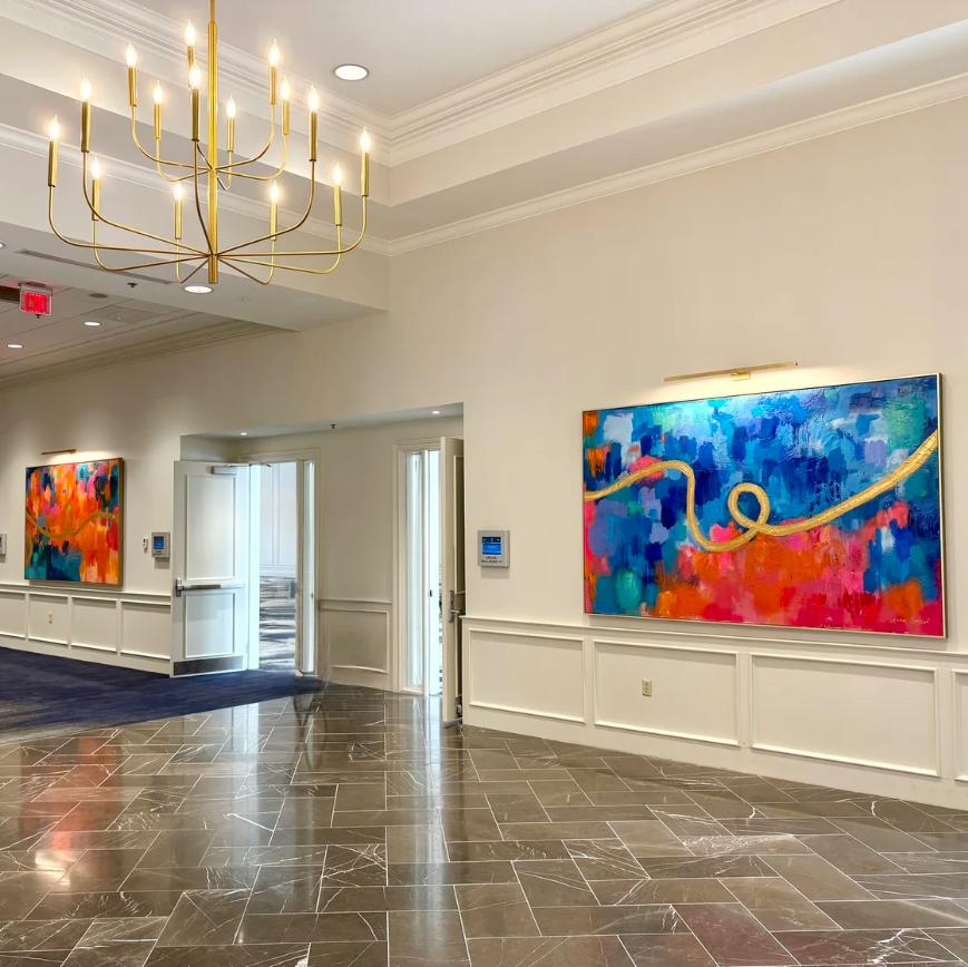 A large abstract canvas painting with vibrant blue, red, and pink brushstrokes and gold flowing lines, displayed on a white wall in a modern hotel lobby with marble floors and a gold chandelier.