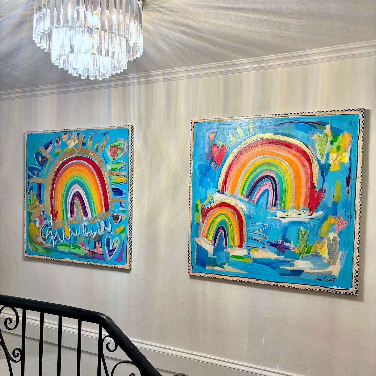 Two colorful framed paintings featuring vibrant rainbow scenes hanging on a white paneled wall above a black metal staircase railing, with a crystal chandelier visible in the upper left.
