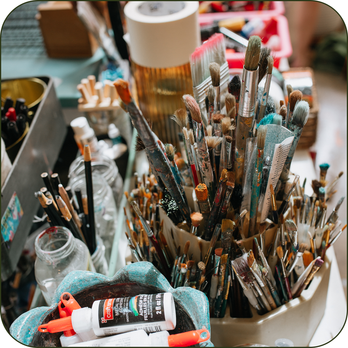A close-up, slightly high-angle view of an artist's stationery setup, featuring a large collection of paint brushes in various sizes and colors held in a beige cup, alongside other art supplies like a tube of acrylic paint, a small bottle, and a teal flower-shaped holder.