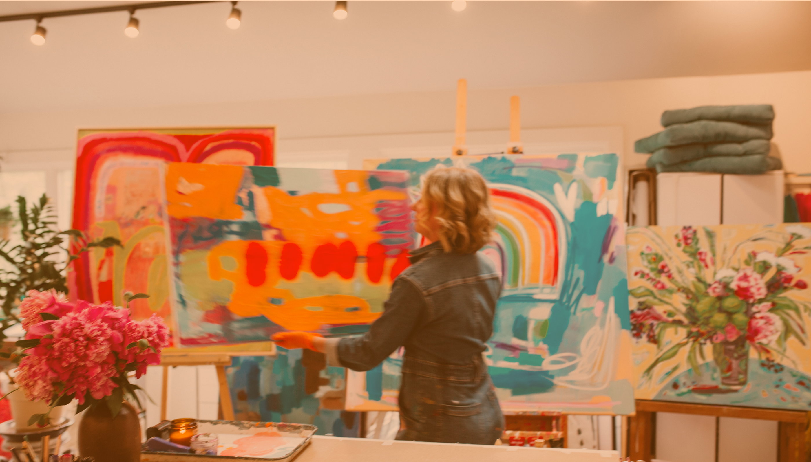 A woman painting in a warm, brightly lit studio, surrounded by colorful canvases on easels, flowers, and folded textiles.