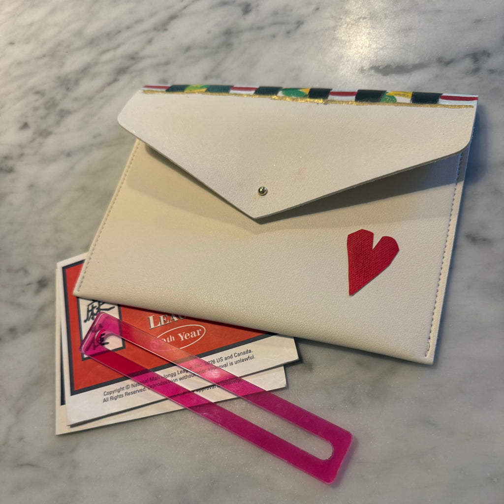 A cream-colored envelope-style card holder wallet with a red heart on the front, shown on a white marble surface with a pink plastic tag and printed cards underneath.