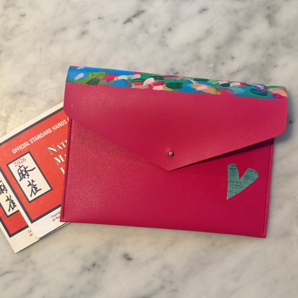 A bright pink envelope-style wallet or card holder with a flap closure and a colorful floral-patterned side panel, shown on a white marble surface with a few cards partially tucked inside.