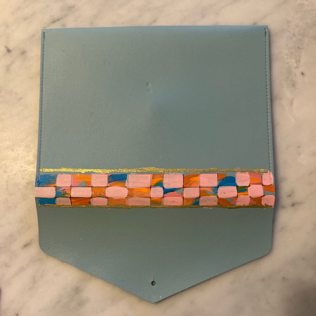 A light blue leather pouch with a colorful geometric patterned band across the middle, shown on a white marble surface with a hand visible at the bottom.