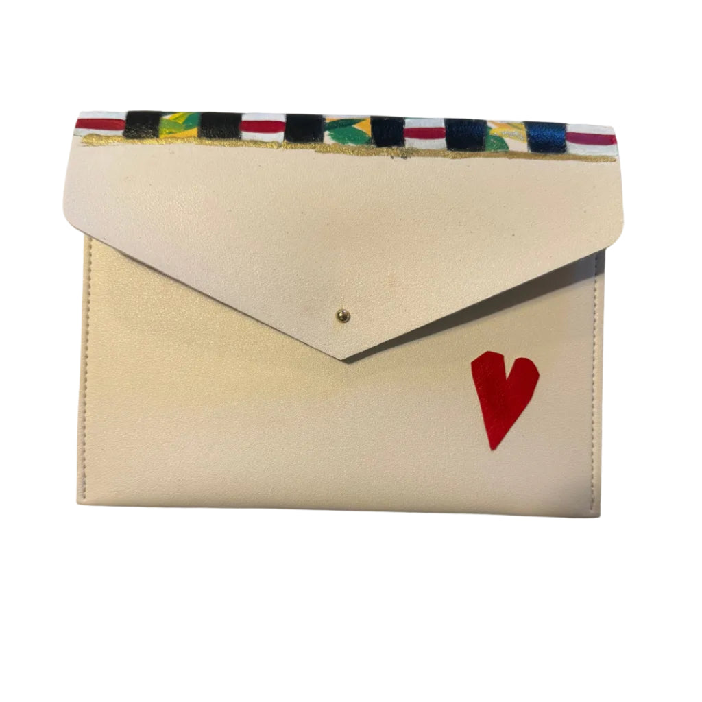 A cream-colored leather envelope-style clutch wallet with a small red heart on the front, featuring a colorful patterned ribbon bookmark or strap across the top, resting on a gray marble surface.