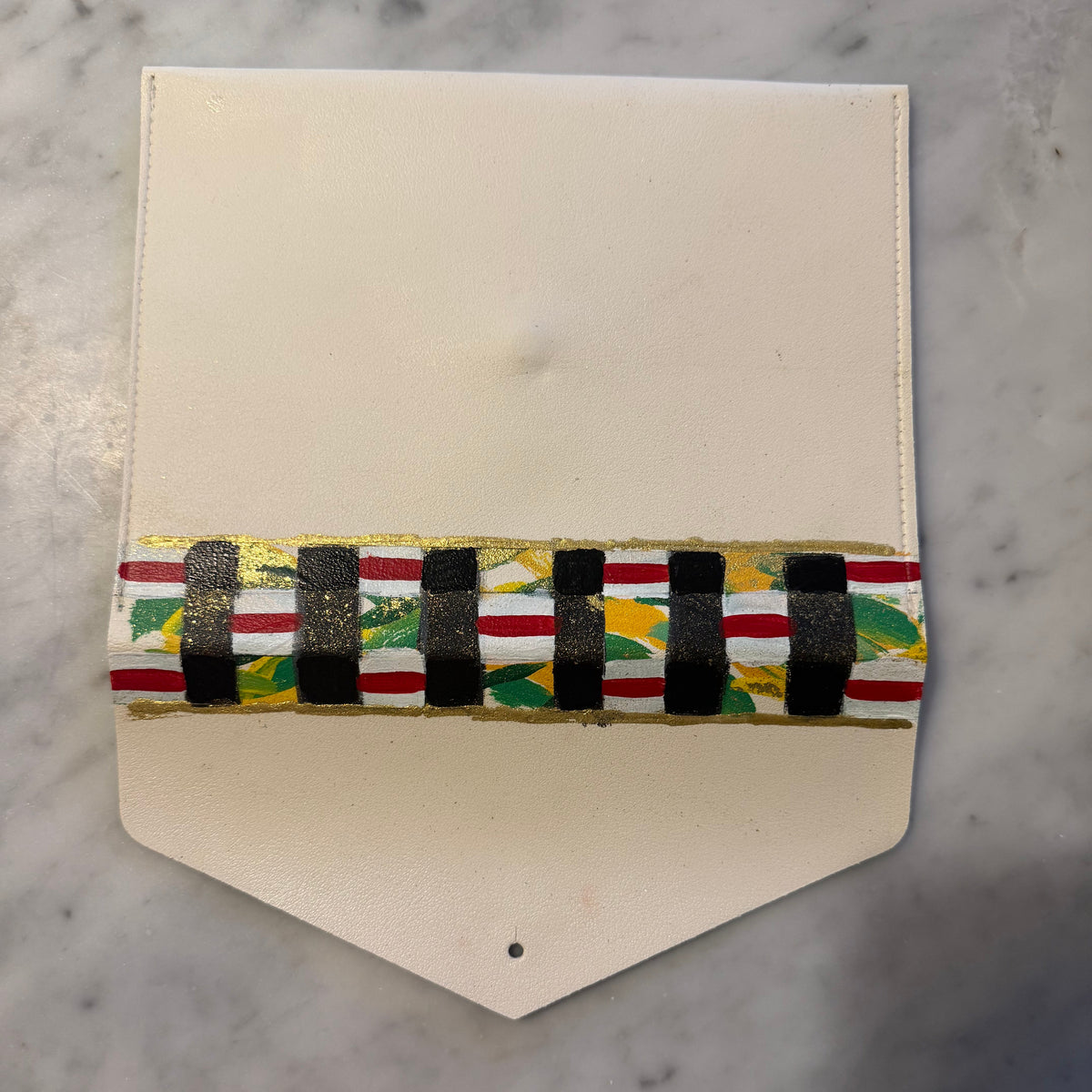 A cream-colored leather pouch with a decorative horizontal band featuring black, red, white, green, and gold patterns, shown on a marble surface with a hand visible at the bottom left.