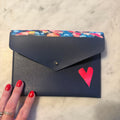A navy blue envelope-style clutch wallet with a colorful floral patterned top border, a small gold snap button, and a bright pink heart on the front, held by a hand with red nail polish on a light marble surface.