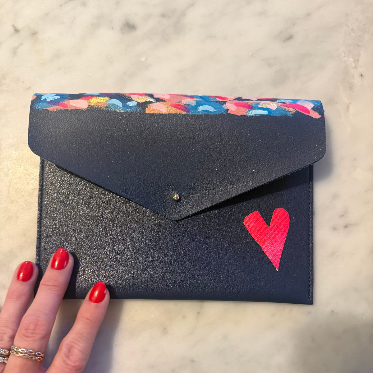 A navy blue envelope-style clutch wallet with a colorful floral patterned top border, a small gold snap button, and a bright pink heart on the front, held by a hand with red nail polish on a light marble surface.