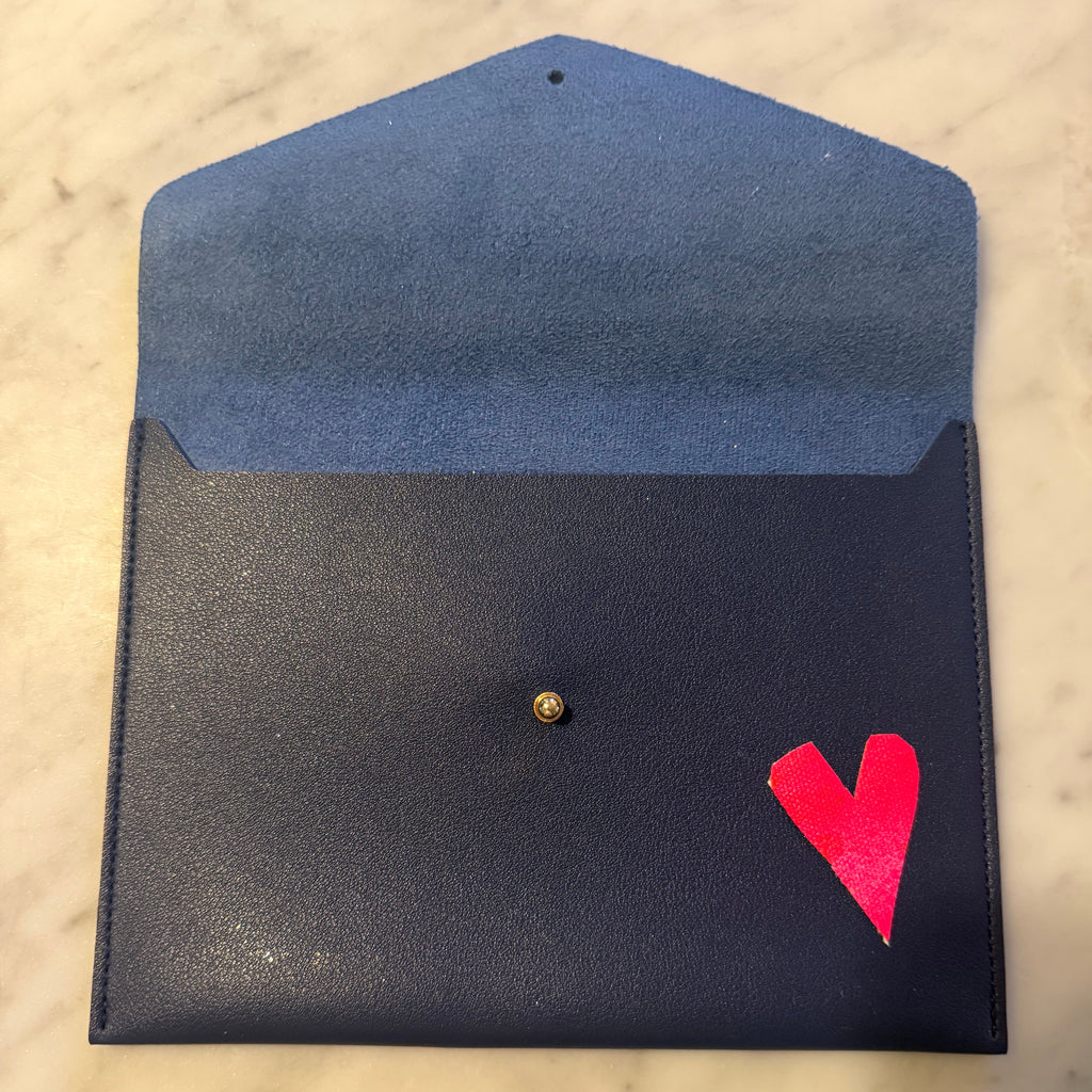 A dark navy blue envelope-style pouch with a lighter blue interior flap, a small gold snap button, and a bright pink heart detail on the front, shown on a light marble surface.