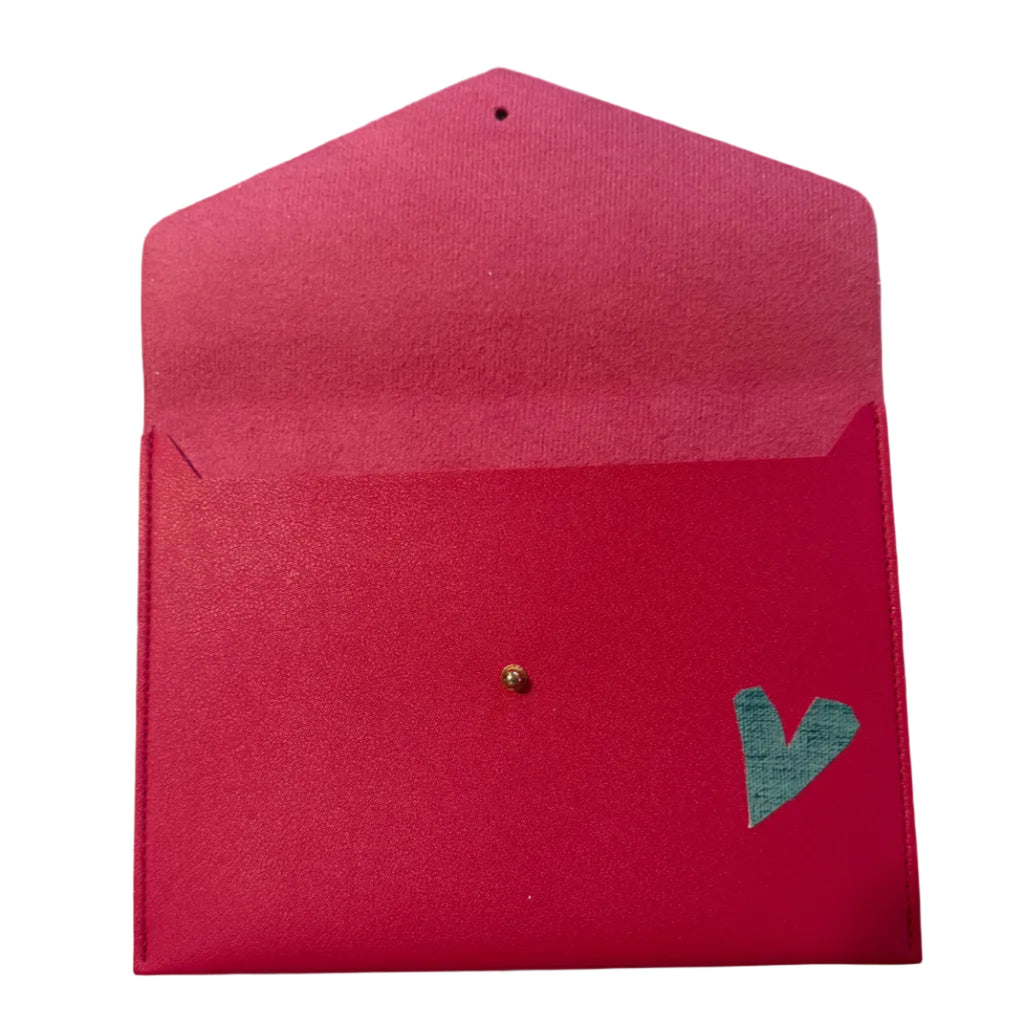 A bright pink textured paper envelope with a small gold stud and a green heart-shaped embellishment, shown on a white marble surface.