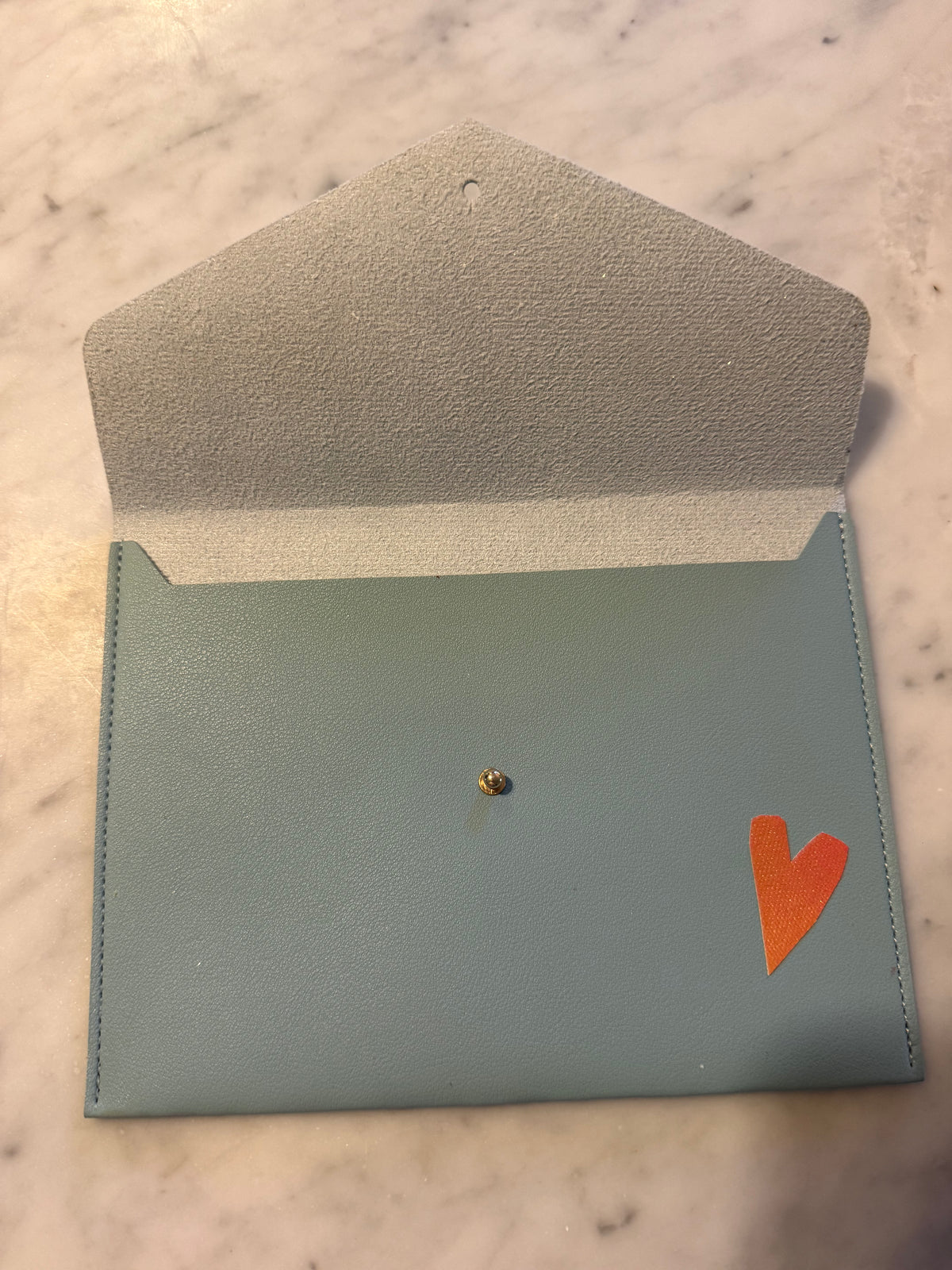 A light blue leather card holder or envelope-style wallet with a small orange heart on the front, photographed on a light marble surface.