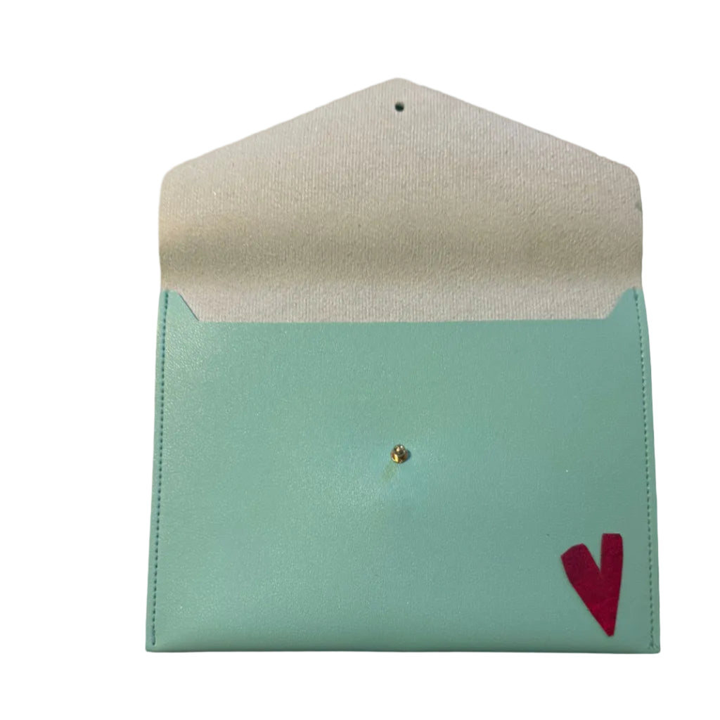 A mint green faux leather envelope-style pouch with a white textured interior lining, a small gold rhinestone clasp, and a small red heart detail on the front, shown on a marble surface with a hand visible in the corner.