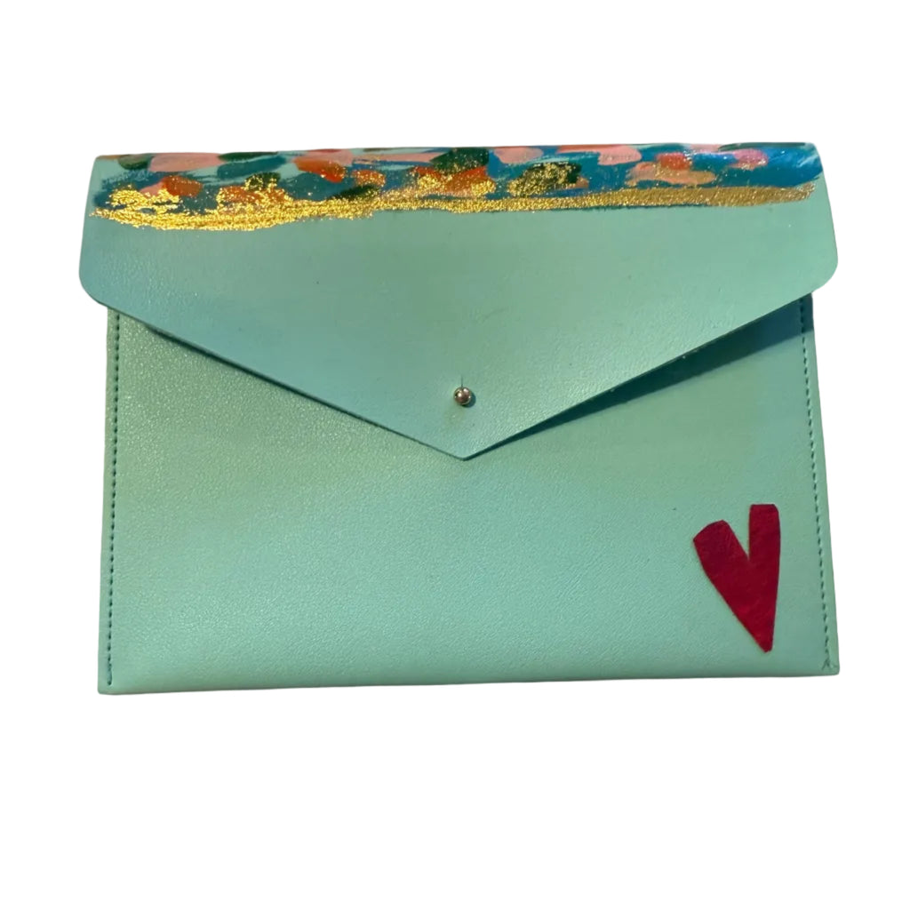 A light blue envelope-style wallet or clutch with a colorful floral patterned trim along the top flap, a small silver snap button, and a red heart detail on the front. A hand with red-painted nails rests near the bottom left on a light marble surface.