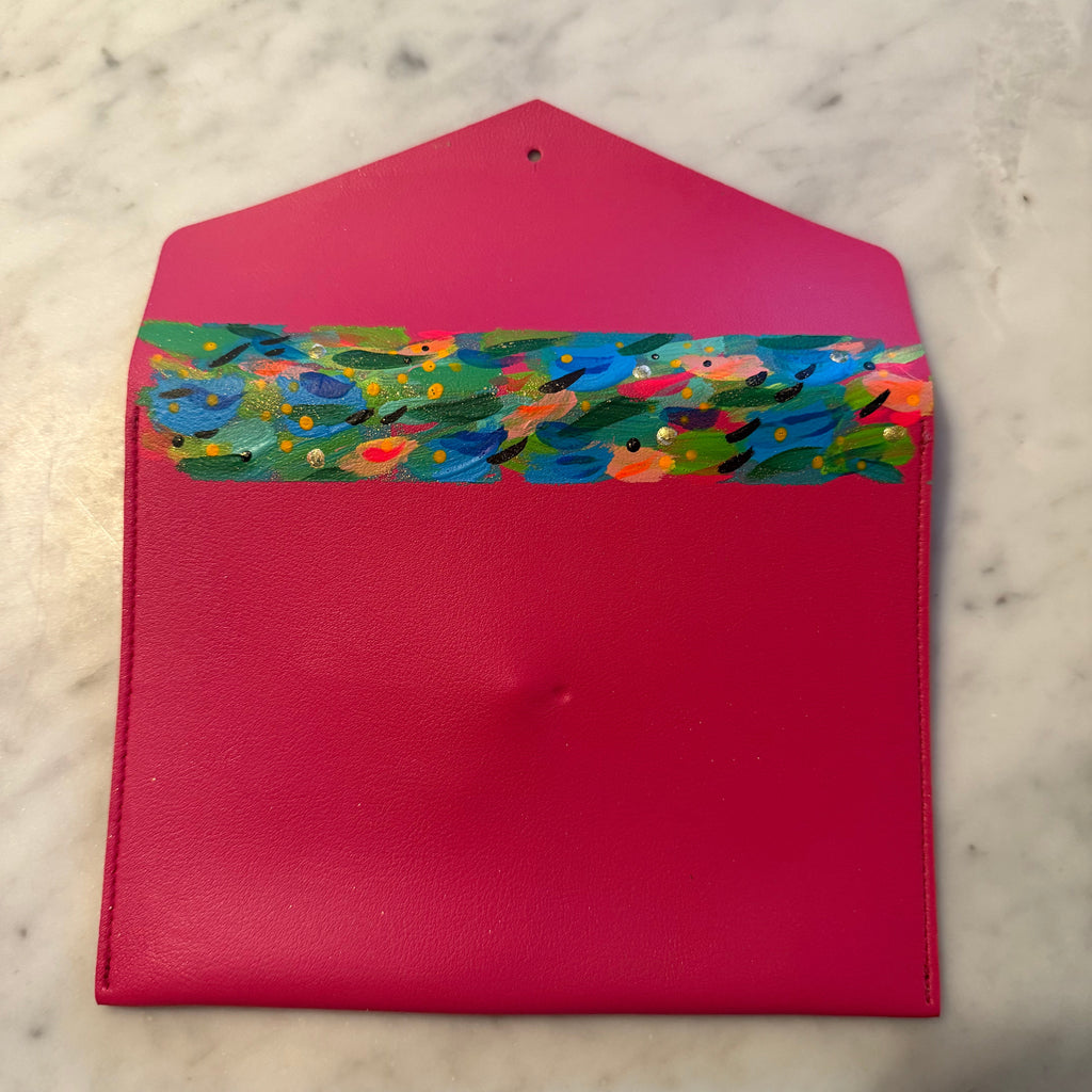 A bright pink faux-leather envelope-style pouch with a colorful hand-painted floral pattern across the top flap, displayed on a light marble surface.