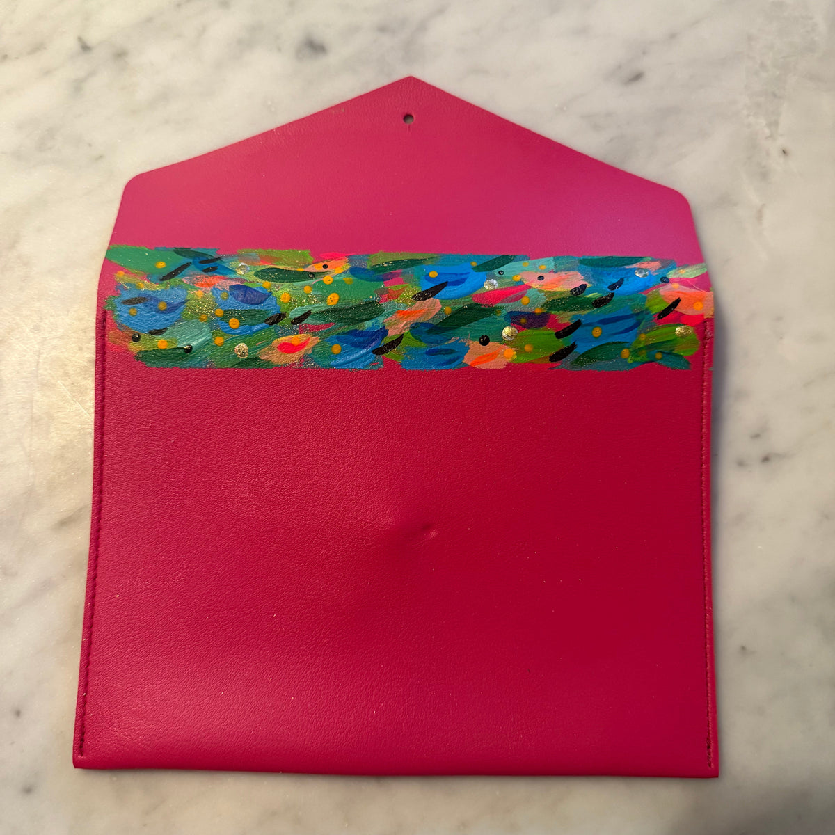 A bright pink faux-leather envelope-style pouch with a colorful hand-painted floral pattern across the top flap, displayed on a light marble surface.
