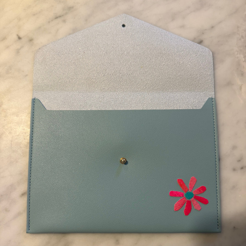 A light blue envelope-style pouch with a white textured interior, a small gold button closure, and a pink flower applique on the front, shown on a marble surface with a hand visible at the bottom.