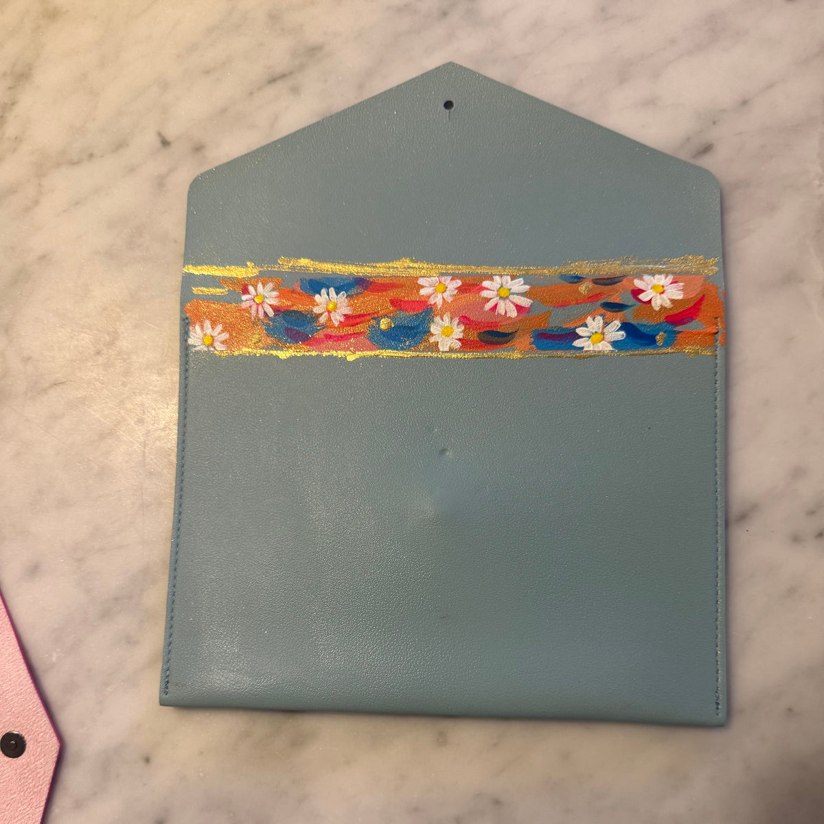 A light blue faux-leather envelope-style clutch wallet with a colorful hand-painted floral stripe across the front, resting on a white marble surface.