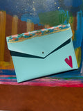 A light blue envelope-style clutch bag with a floral patterned top flap, gold trim, and a pink heart detail, displayed against a colorful painted background.