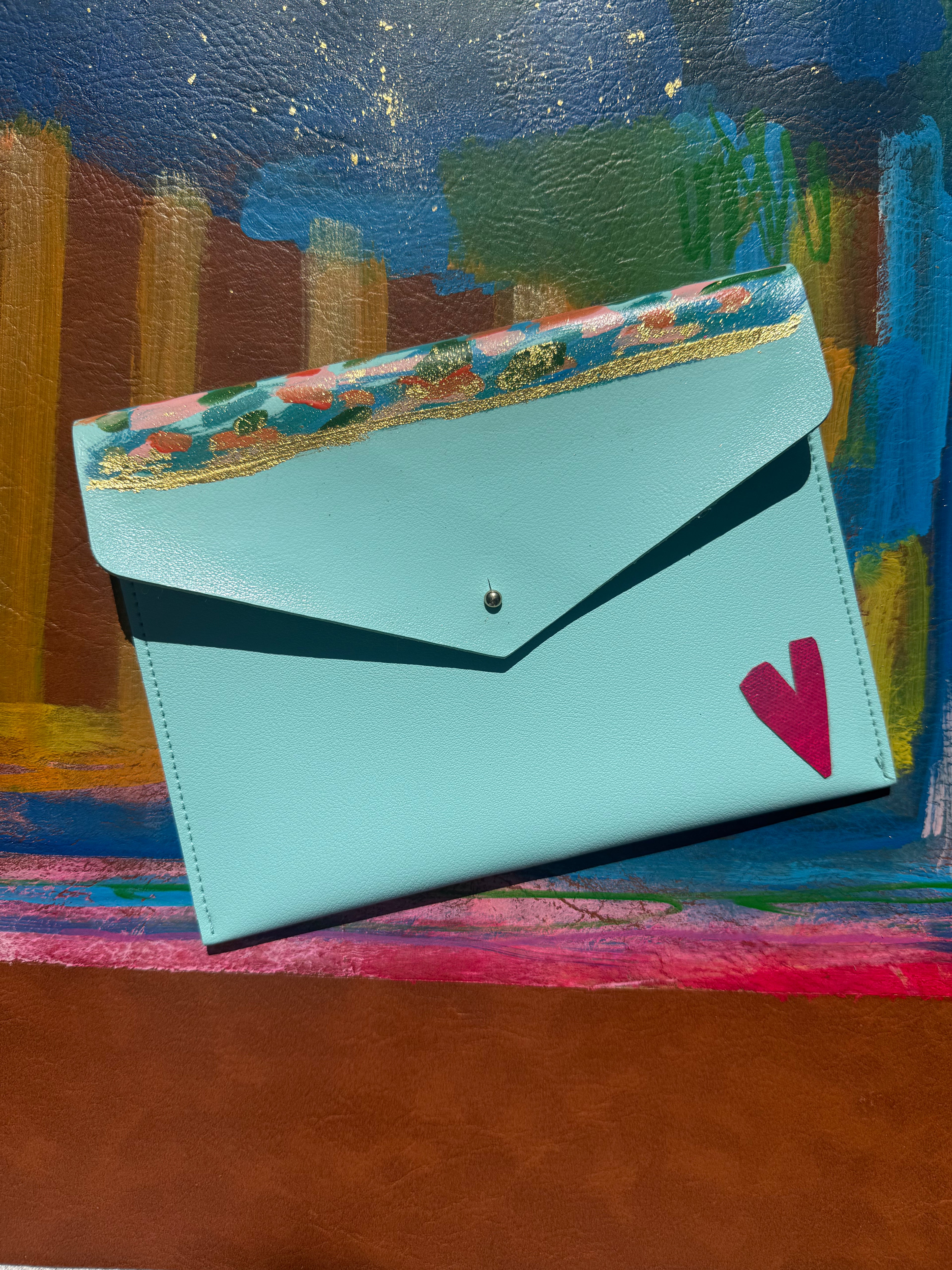 A light blue envelope-style clutch bag with a floral patterned top flap, gold trim, and a pink heart detail, displayed against a colorful painted background.
