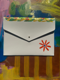 A white envelope-style clutch wallet with a red flower design and colorful patterned border, displayed against a colorful abstract painted background.
