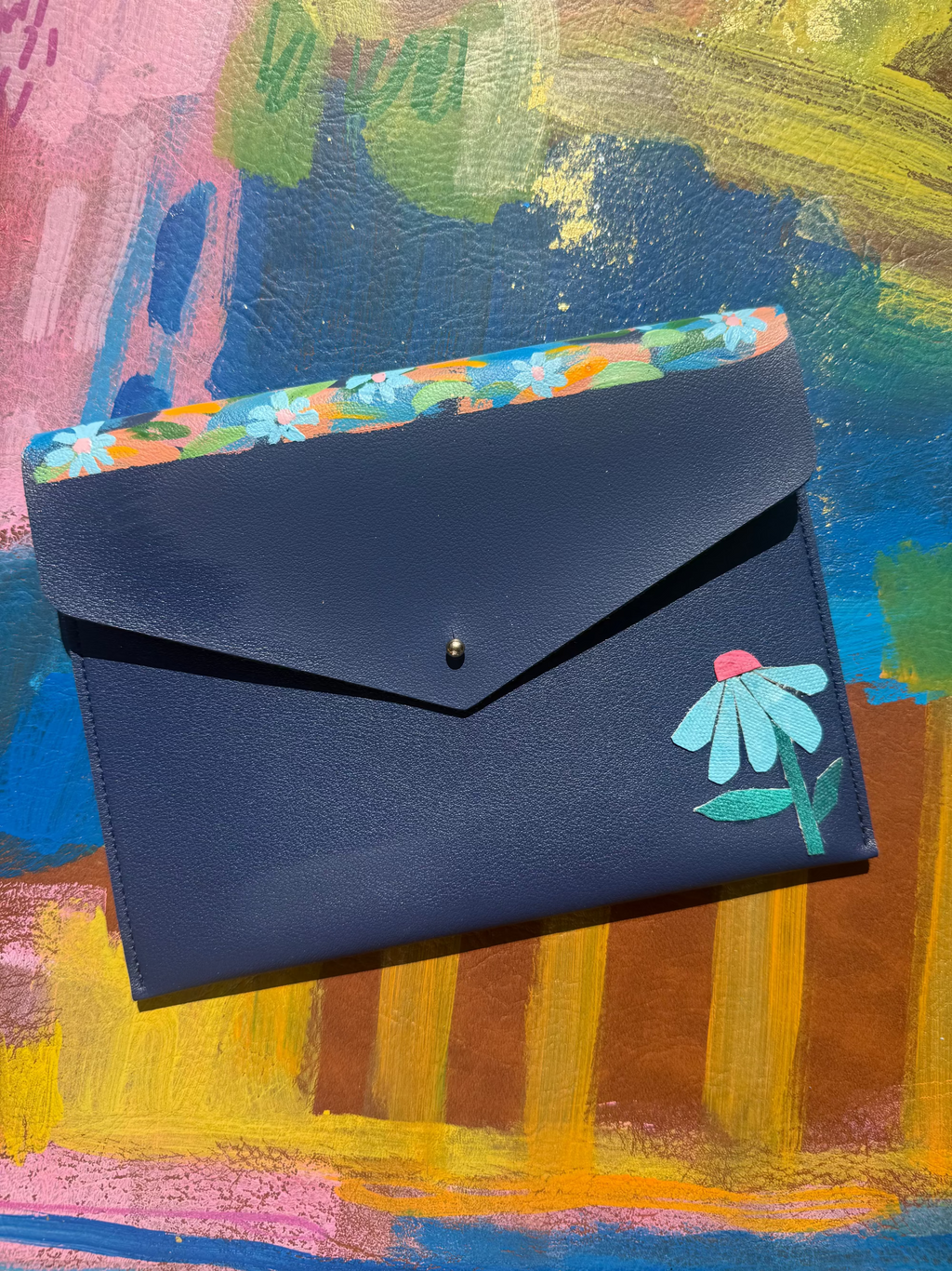 A dark navy blue envelope-style wallet with a small green and pink leaf illustration on the flap, shown open to reveal a colorful floral patterned interior, set against a vibrant abstract painted background.