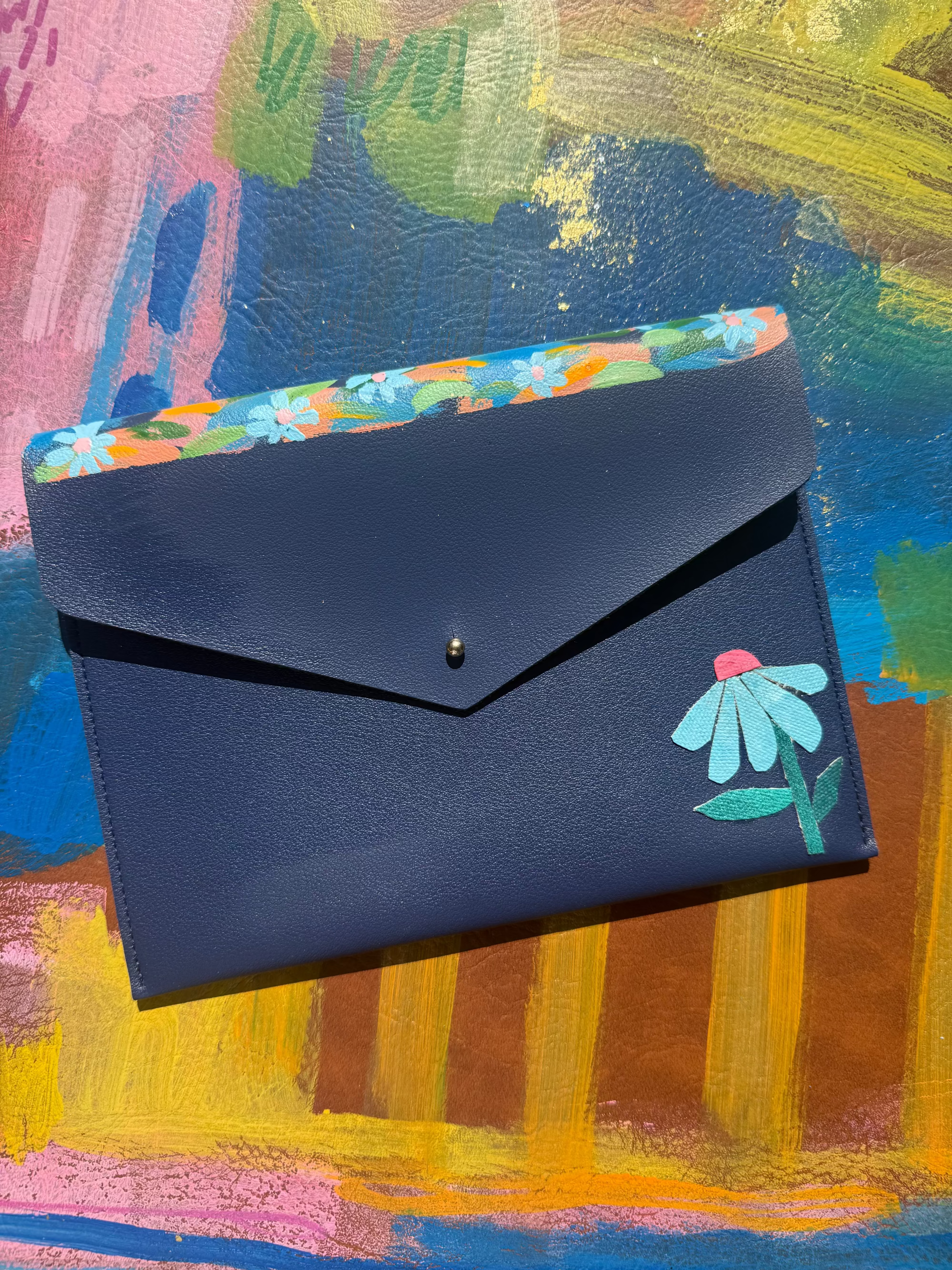 A dark navy blue envelope-style wallet with a small green and pink leaf illustration on the flap, shown open to reveal a colorful floral patterned interior, set against a vibrant abstract painted background.