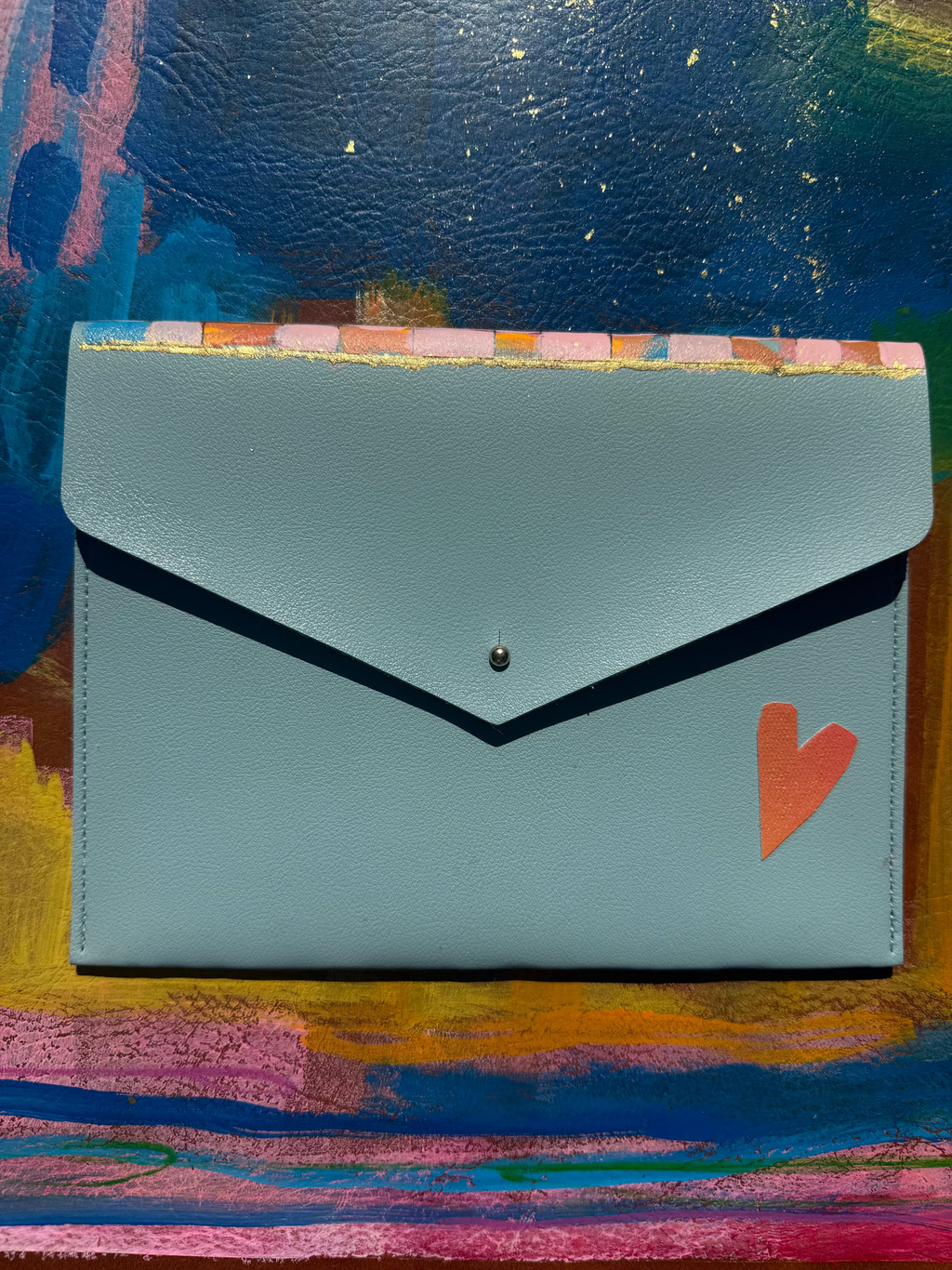 A light blue leather envelope-style wallet with a small orange heart on the left and a colorful striped border along the bottom, displayed on a vibrant painted background.