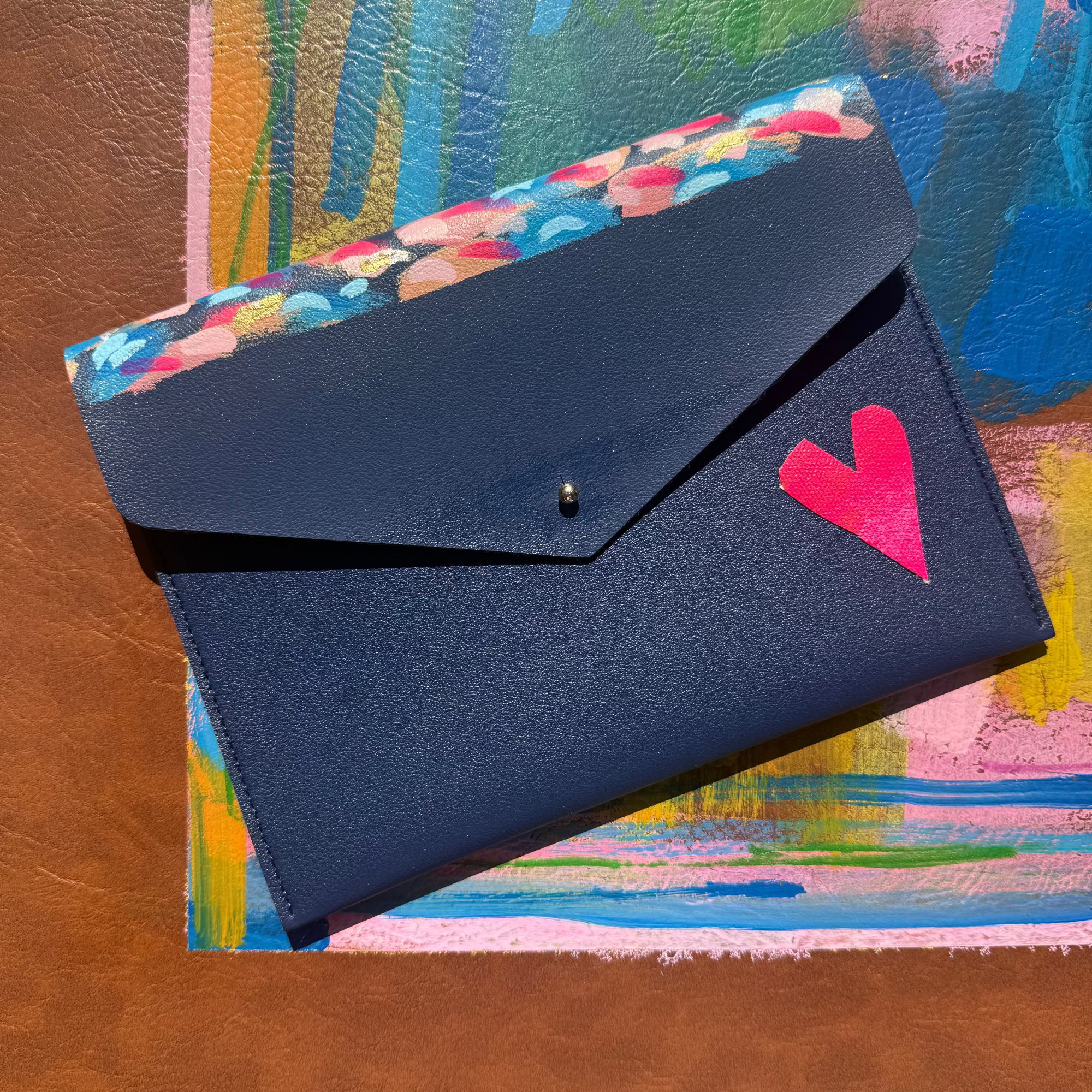 A dark navy blue envelope-style wallet or clutch with a bright pink heart accent, placed diagonally on top of a colorful abstract painting against a brown textured background.