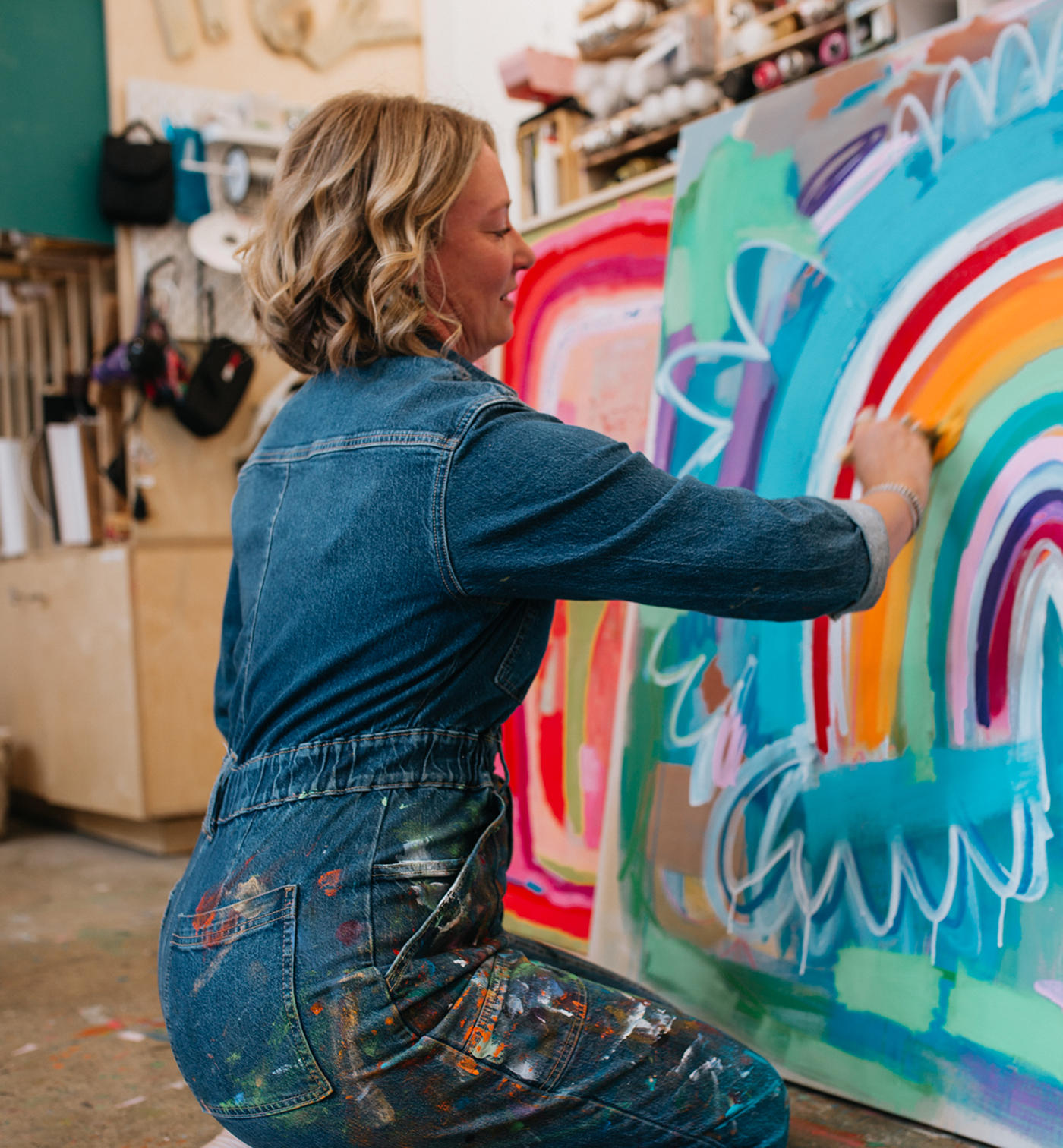 A woman in a blue denim jumpsuit painted with colorful paint is painting a large canvas featuring an abstract rainbow design in a creative studio.