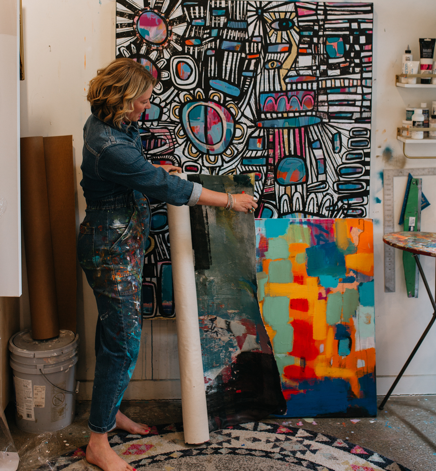 An artist in a paint-splattered denim outfit stands in an art studio, holding a large rolled canvas in front of a colorful abstract painting. Another vibrant painting leans against the wall on the right, and shelves with art supplies are visible in the background.