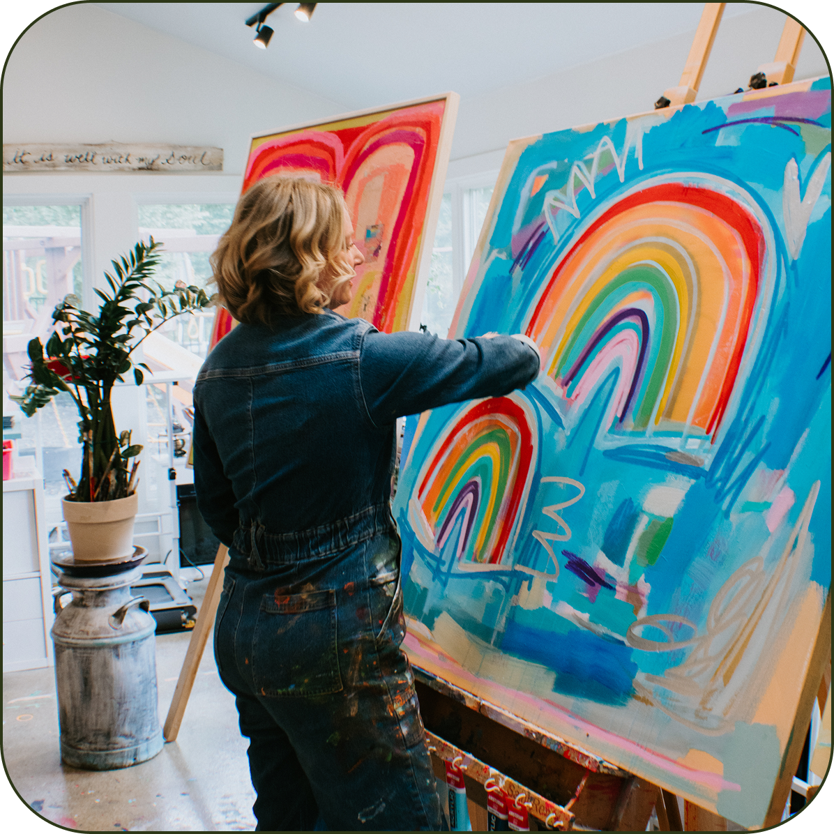 A woman in a blue denim jumpsuit stands at an easel painting a large colorful abstract artwork with rainbow arches on a bright blue background. Another painting is visible in the background inside a bright studio or home space.